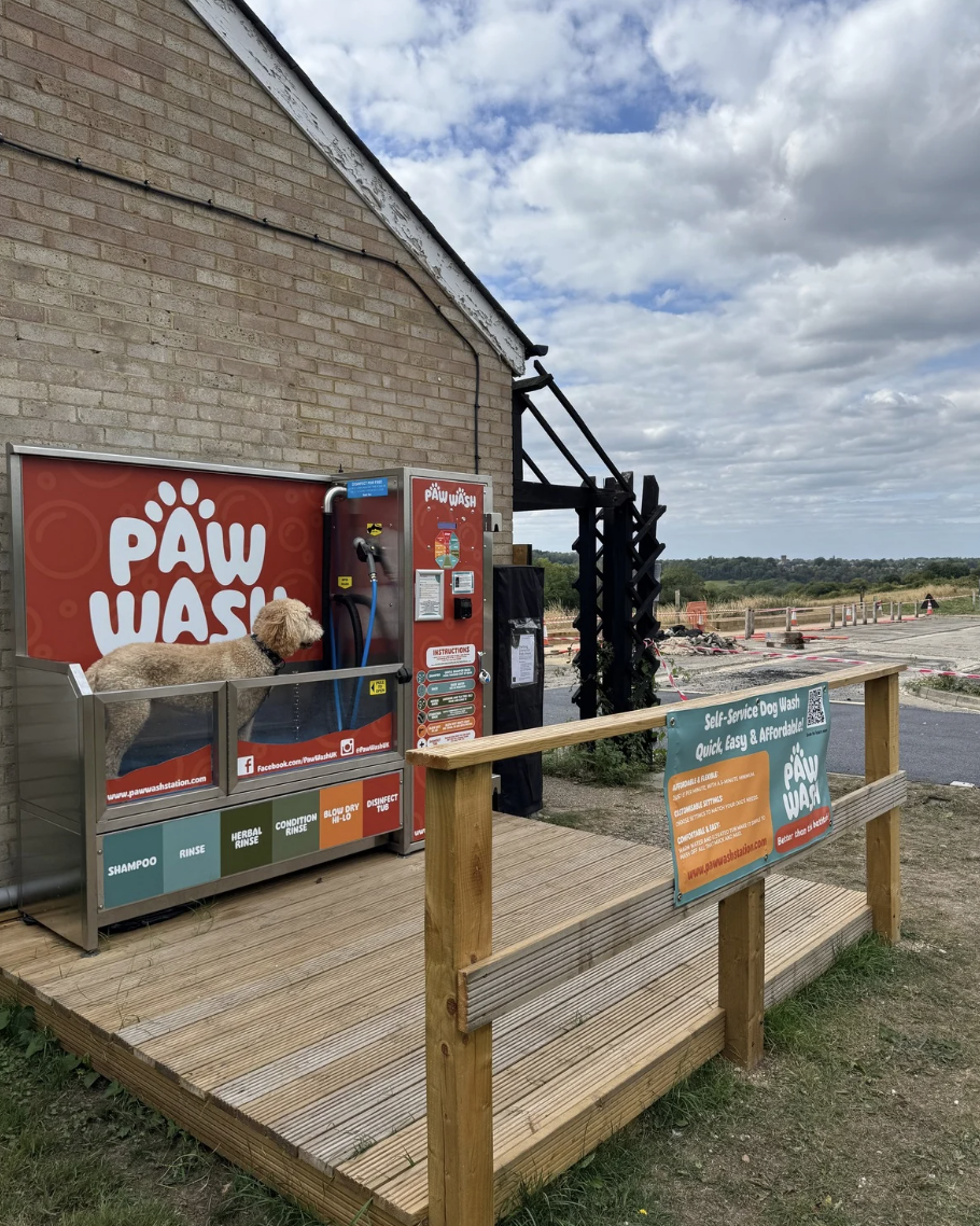 A self-service dog wash station with a dog inside, on a wooden platform outside a brick building. The station has signage with instructions and pricing, and a sign on the fence advertises quick and affordable dog washing services.