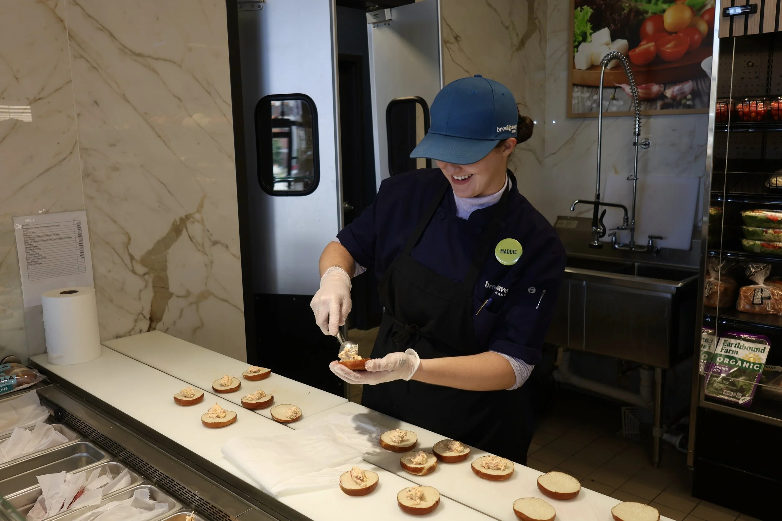 A woman in a blue cap and apron spreads a creamy topping on sliced bread at a food counter, smiling as she works.