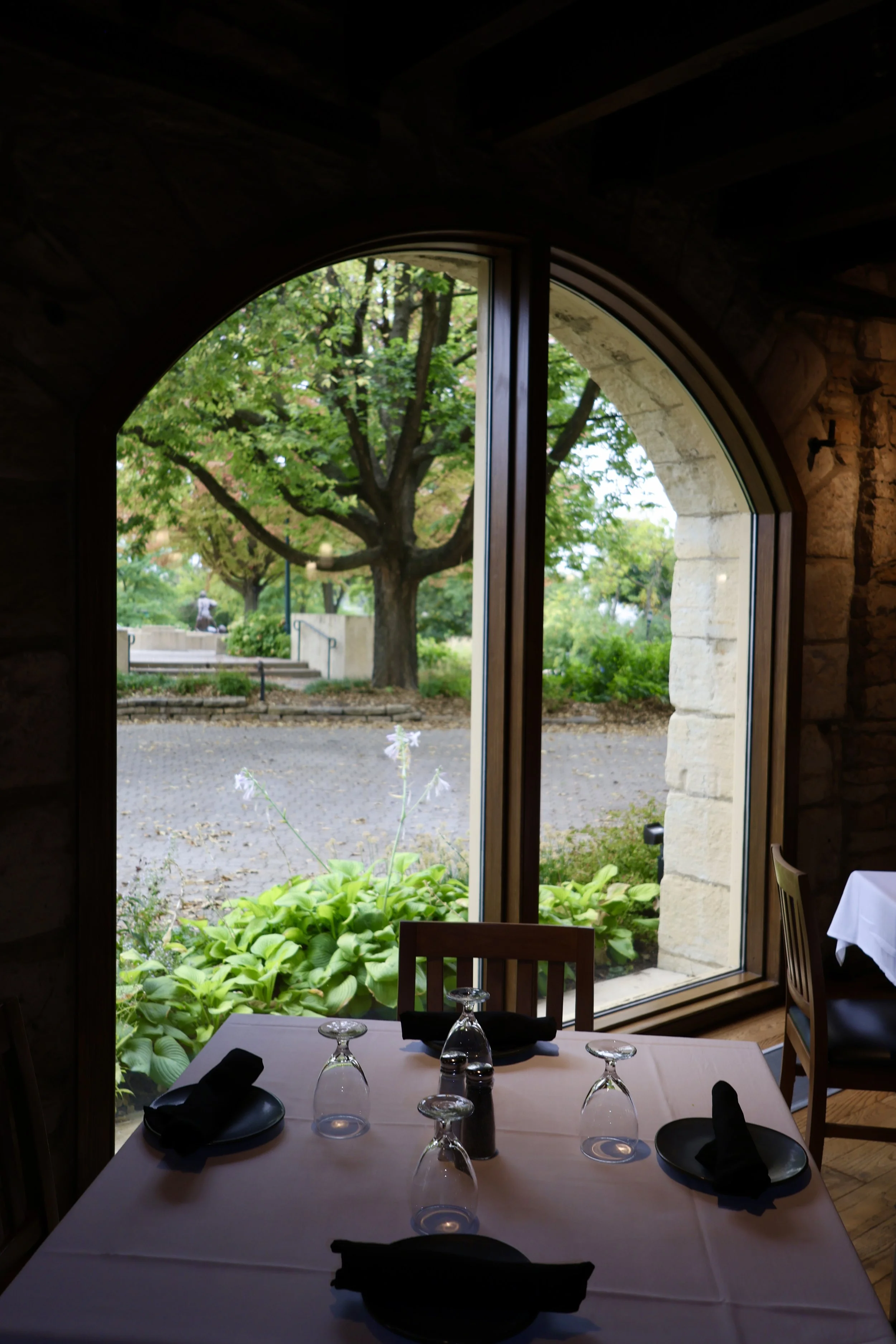 Indoor dining table with black napkins and glasses, overlooking a lush garden through large arched window.