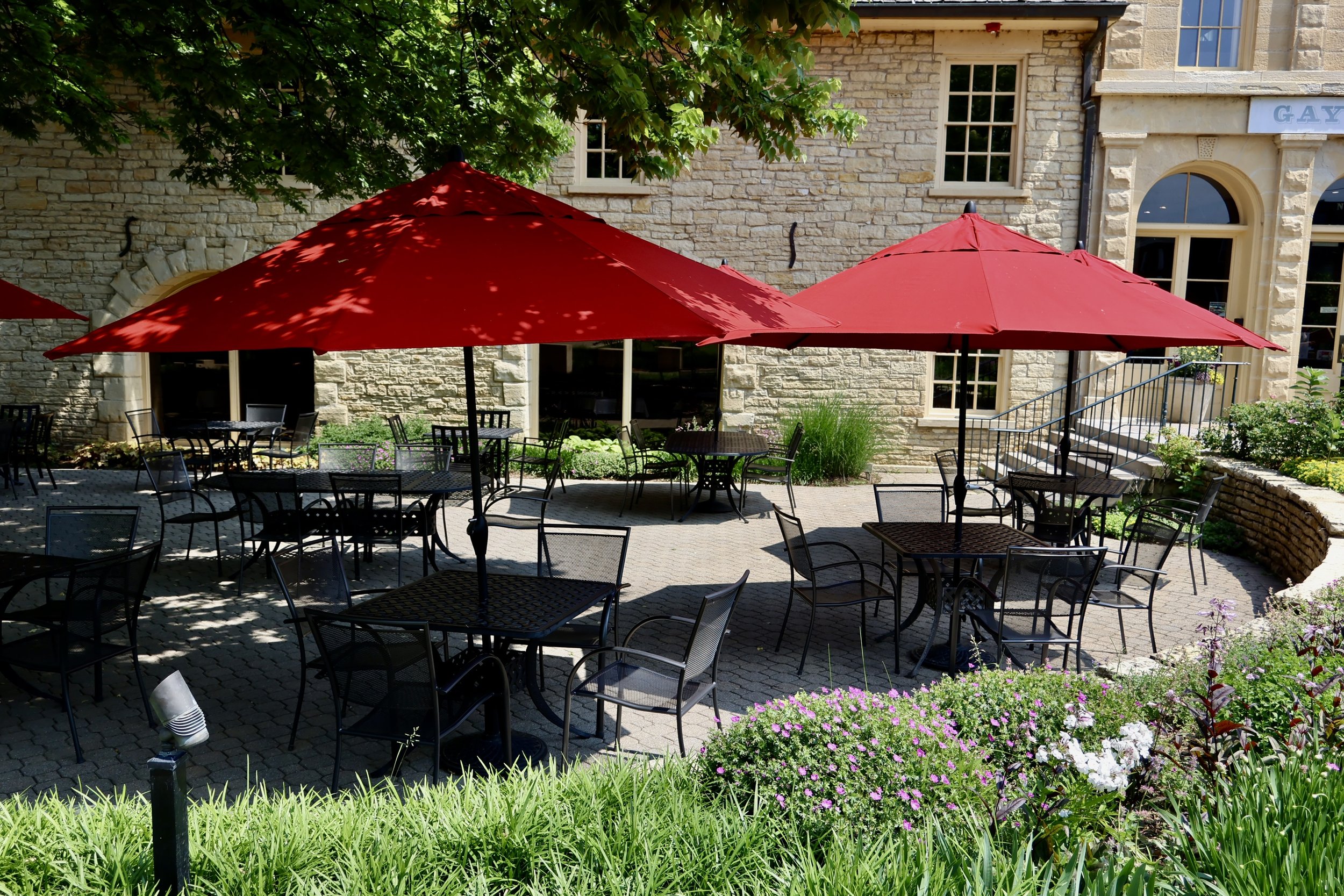Outdoor patio with several black metal tables and chairs, shaded by large red umbrellas, surrounded by greenery and flowers, with a stone building in the background.