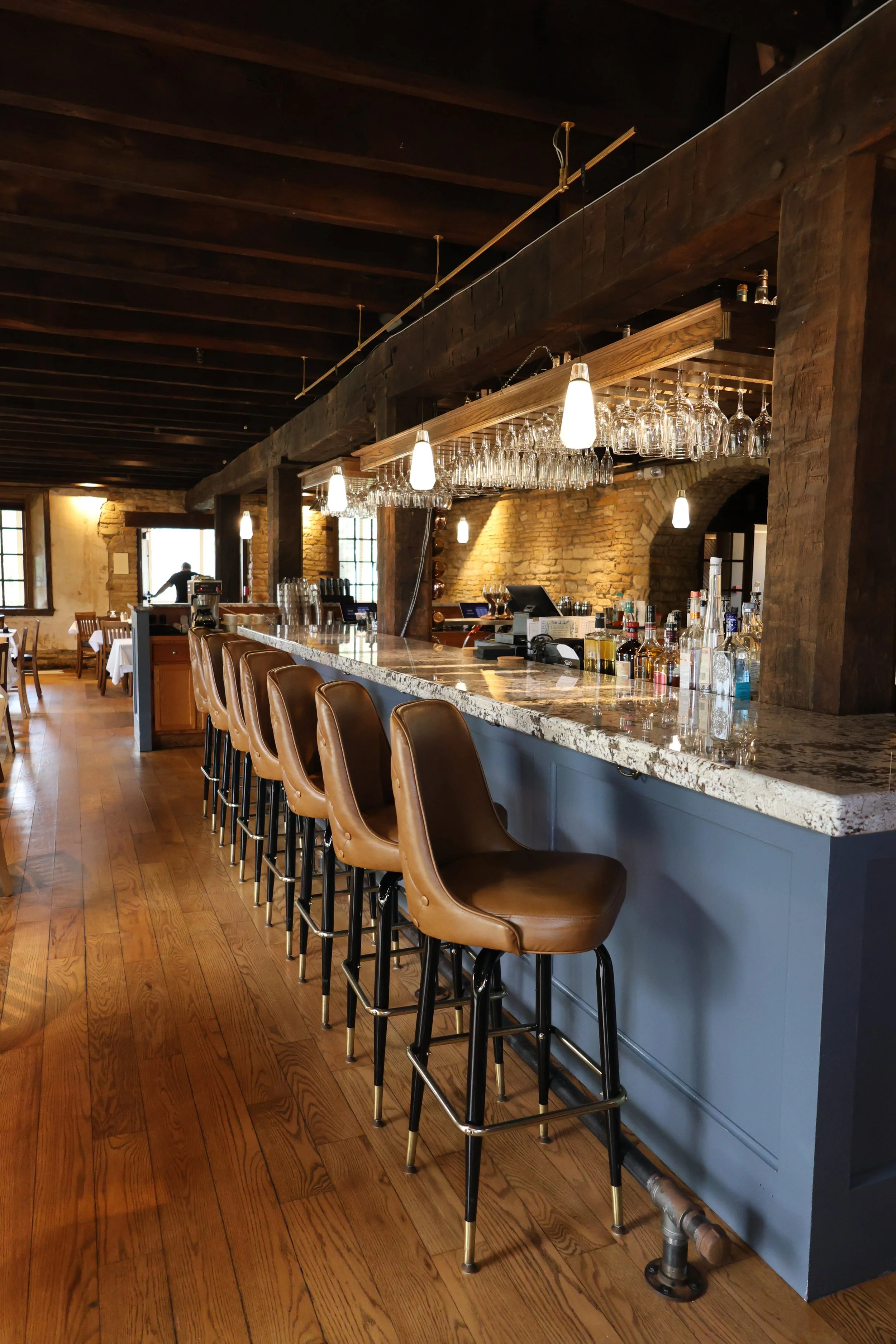 Interior of a rustic bar with brown leather barstools lined up along a marble-topped counter, hanging glasses, and warm ambient lighting.