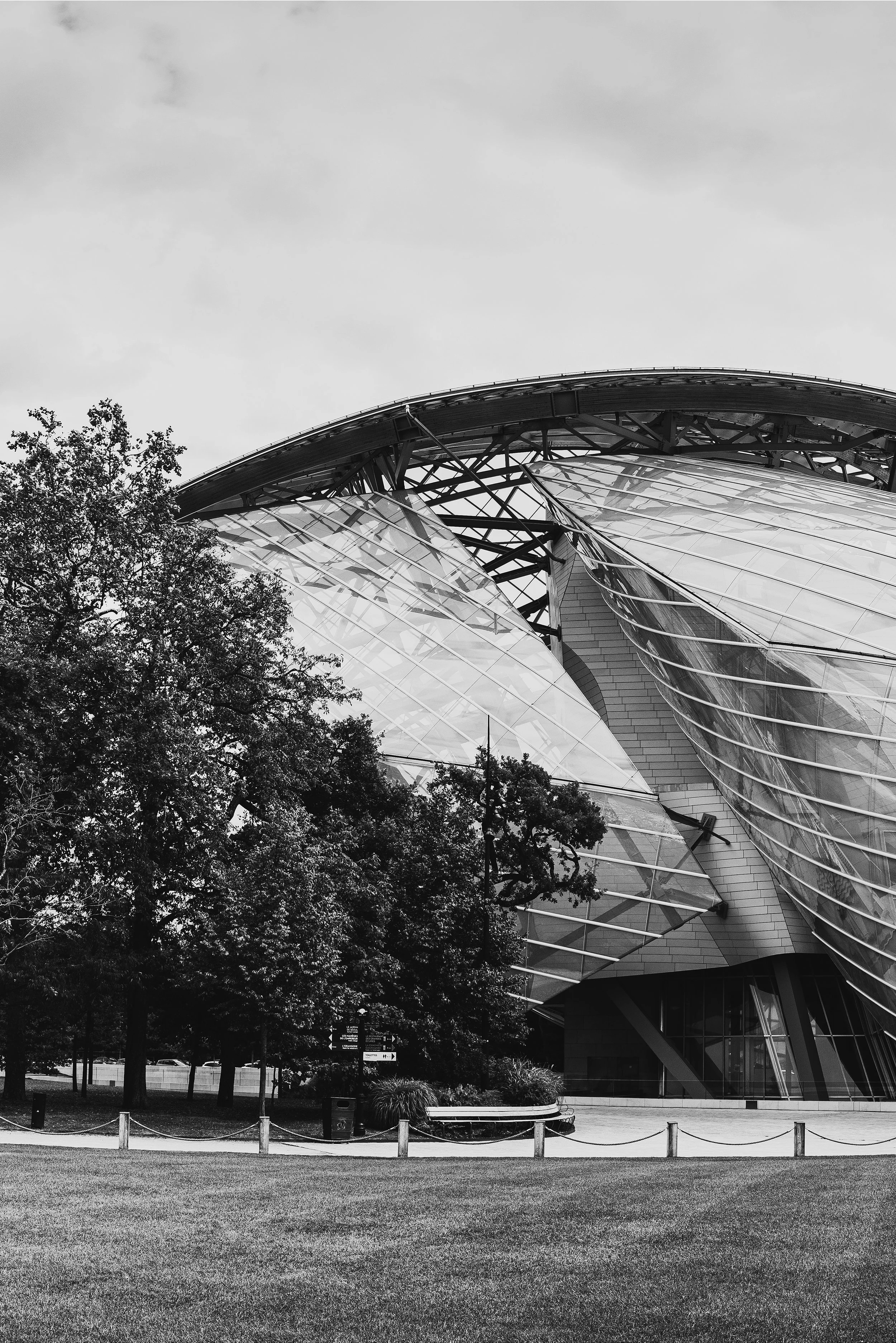A modern architectural building with a curved, glass exterior and metal framework, partially obscured by trees and set against a cloudy sky. Frank Gehry