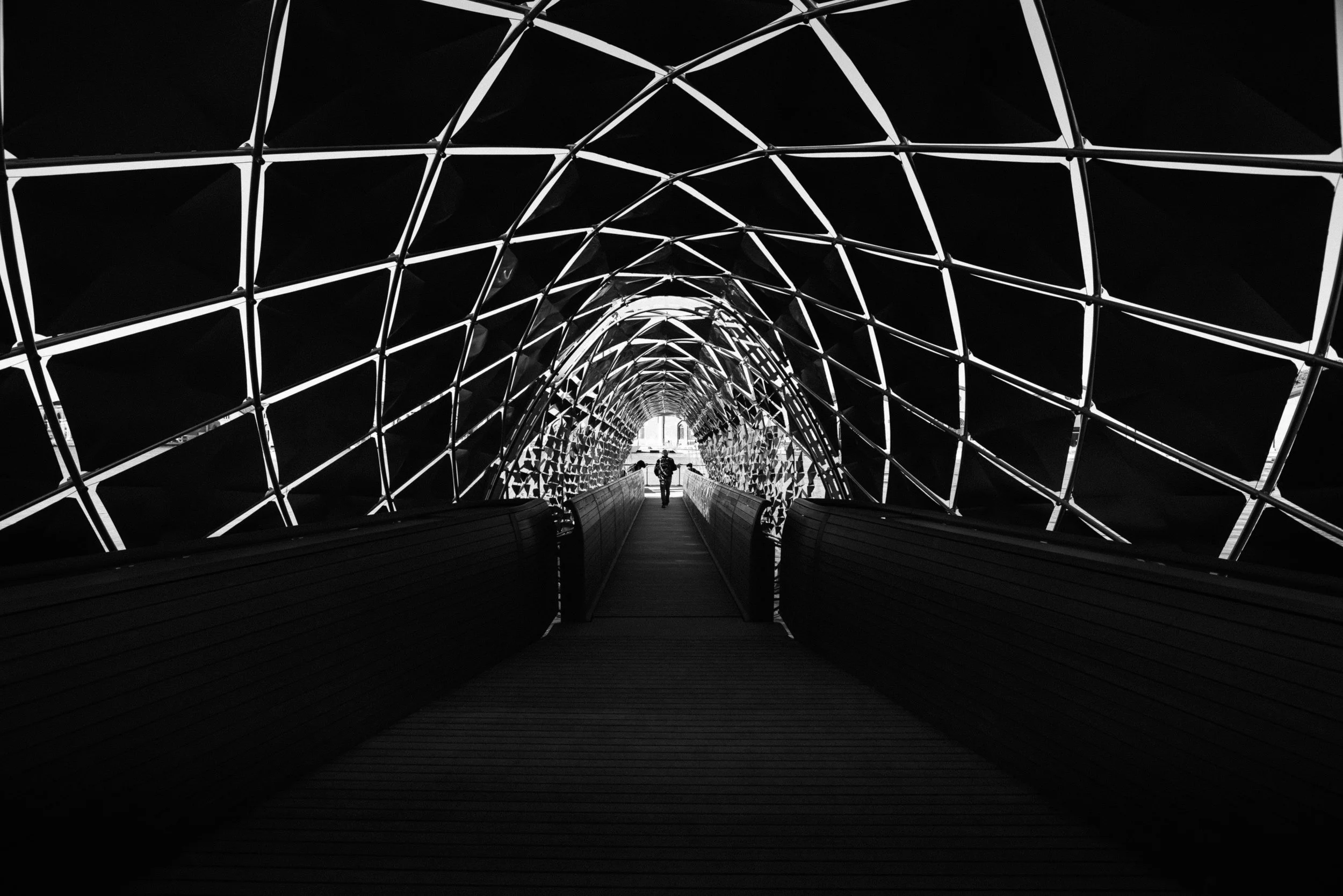 A person walking through a modern, geometric, illuminated tunnel with lattice framework, in black and white. Foster & Partners. Venice Biennale 2025