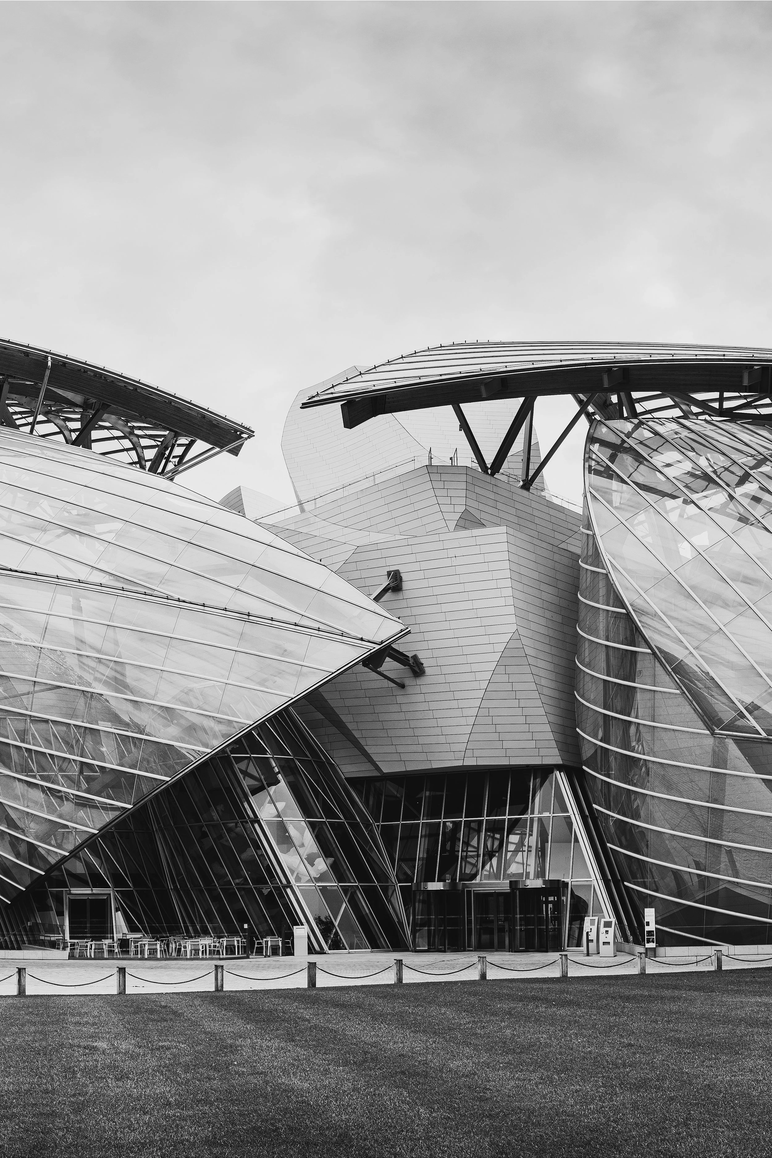 Modern architectural building with glass and metal structures, set against a cloudy sky, in black and white. Frank Gehry