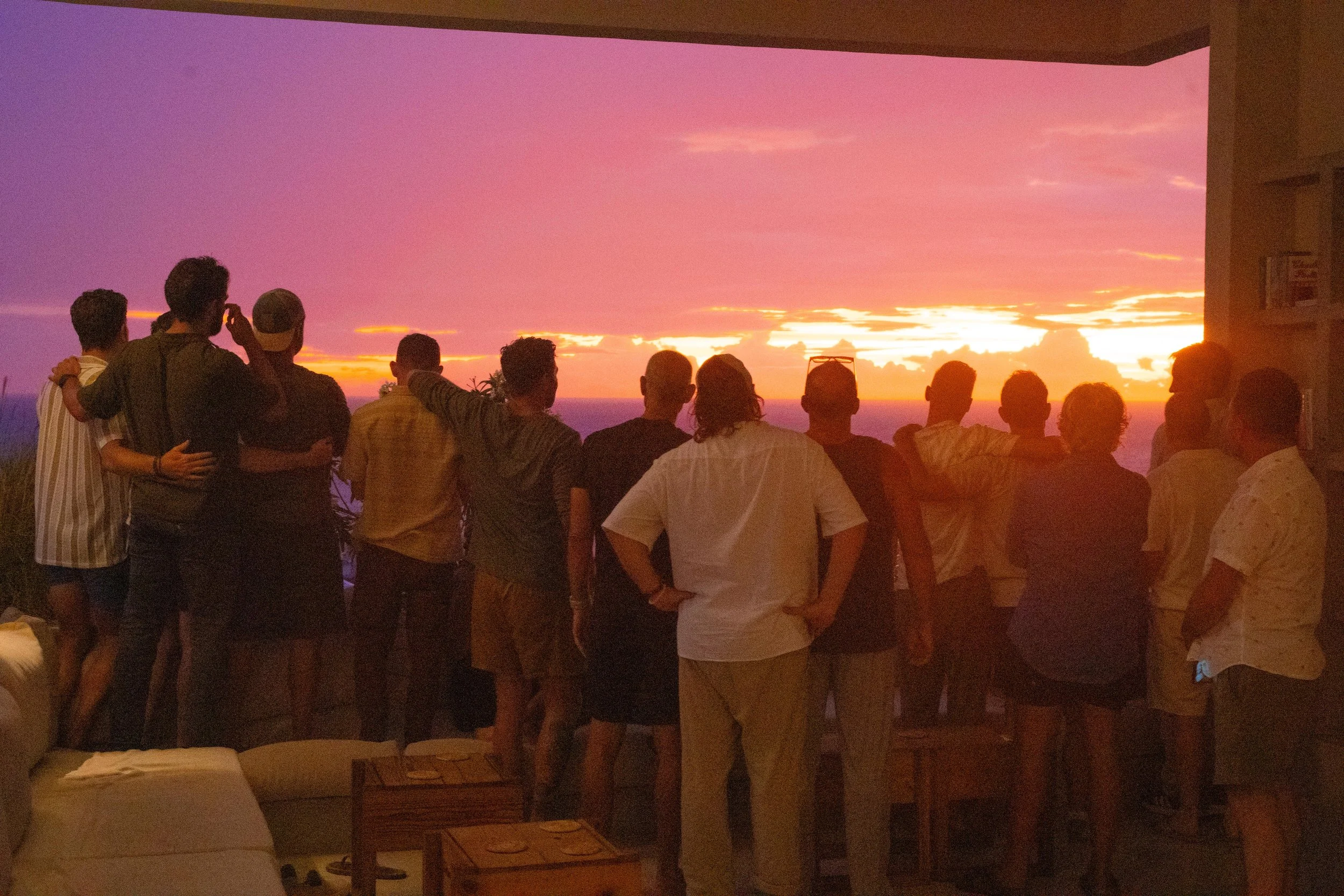 Men standing side by side on a balcony overlooking the ocean at sunset, arm in arm, sharing community and reflection during a somatic wellness retreat in Mexico.