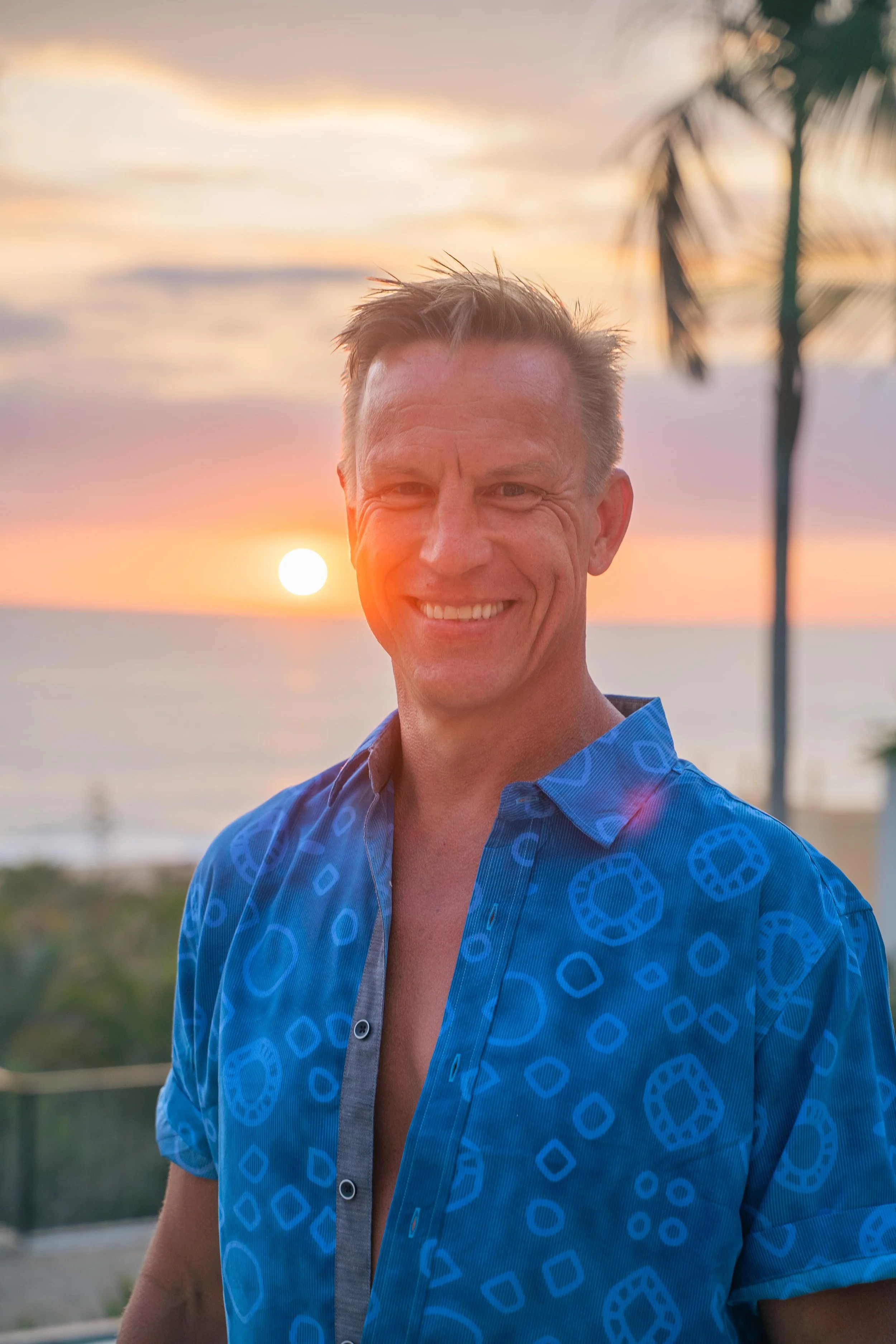 Man smiling in a blue patterned shirt at sunset with the ocean in the background, representing joy, presence, and aliveness at a men’s retreat.