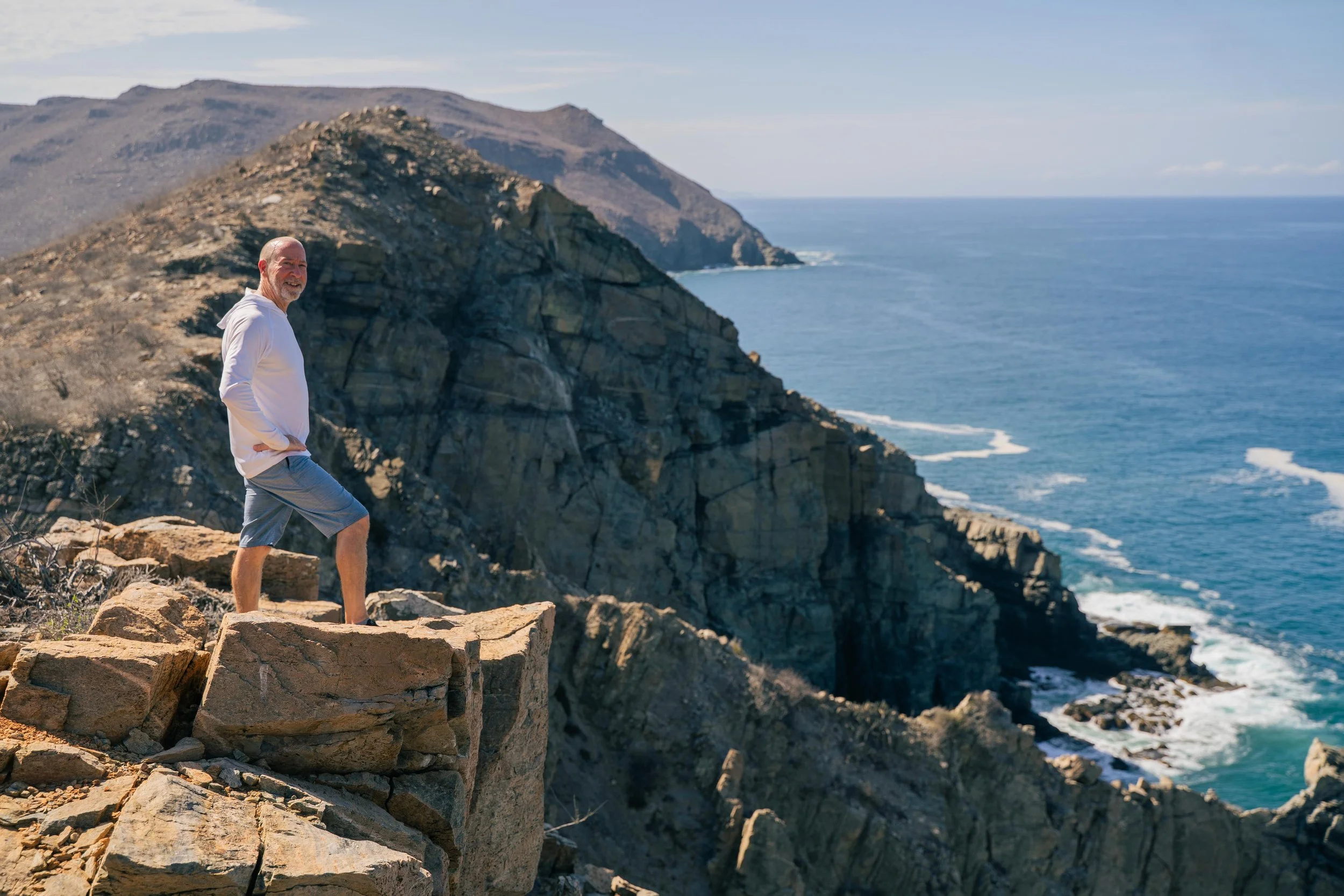 Smiling man standing on a rocky cliff with sweeping ocean views during SHIFT Men’s Retreat, a somatic wellness retreat for men that blends nature, embodiment, and renewal.