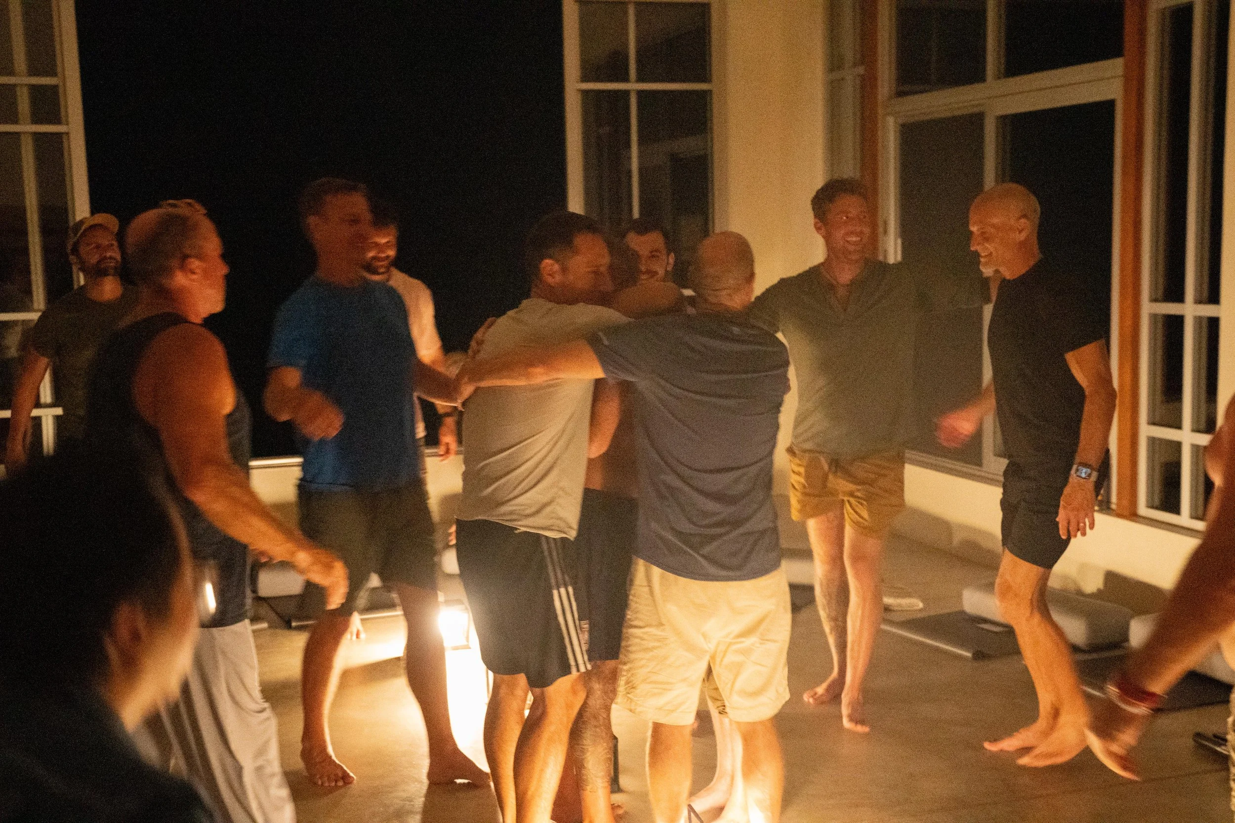 Group of men at a somatic men’s retreat embracing in a circle, barefoot and smiling, during an evening embodiment session focused on connection and nervous system healing.
