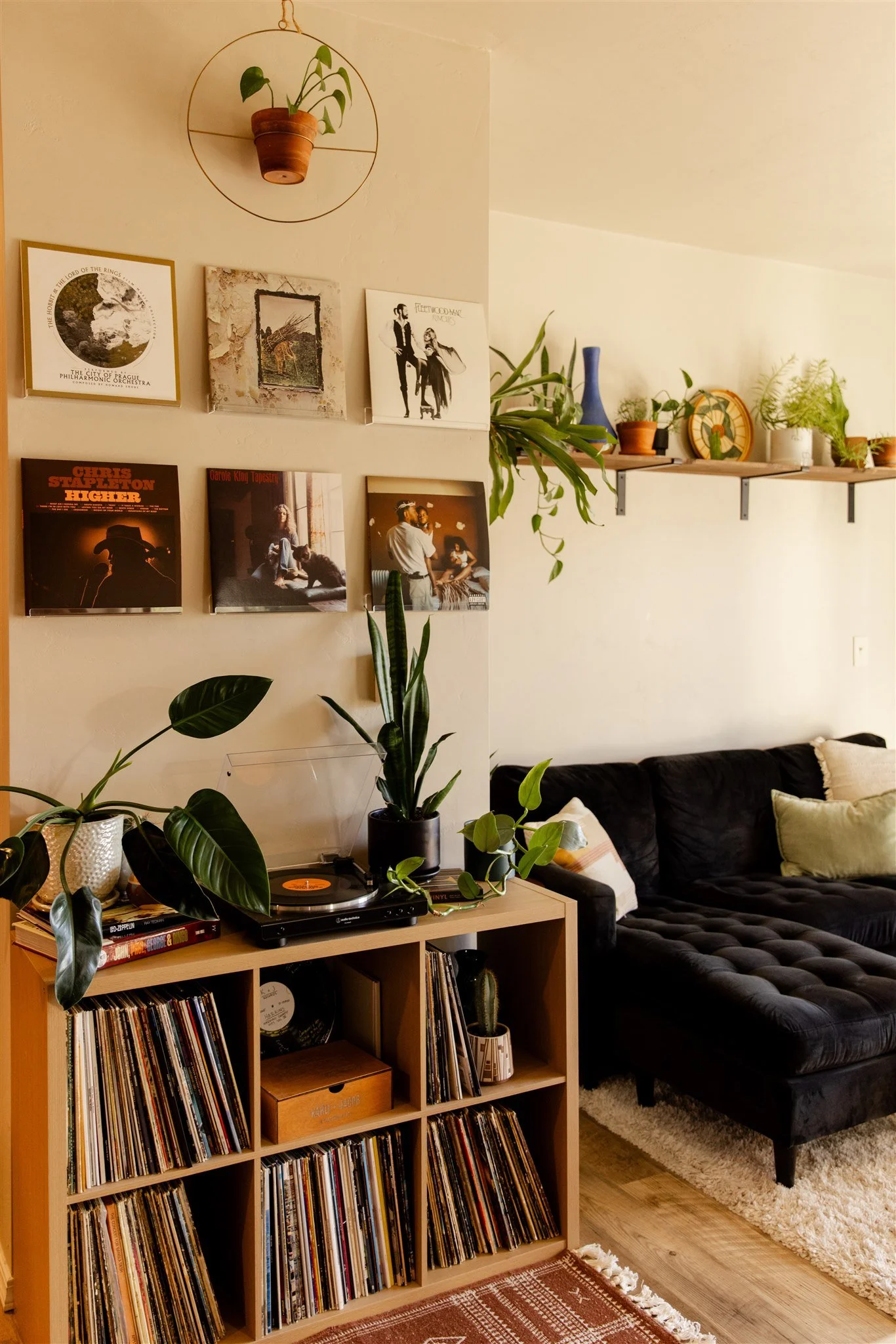 A cozy living room corner featuring a black velvet tufted sofa with pillows, a wooden vinyl record shelf filled with records, potted plants, and a record player. The wall displays a collection of album covers and artwork, with white shelves holding more plants and decorative items.