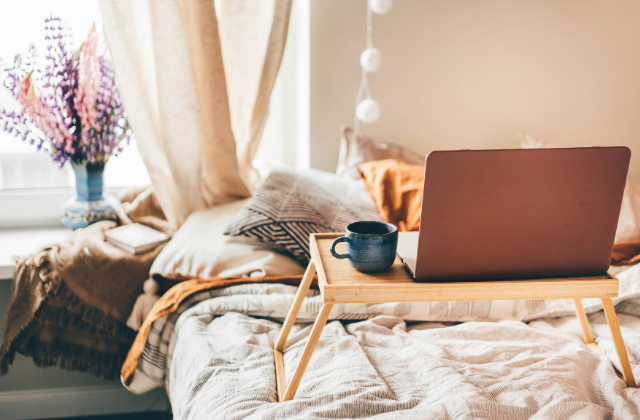 A cozy bedroom with a laptop on a wooden breakfast tray, a cup of coffee, bed with pillows, and a window with curtains and a vase of flowers.