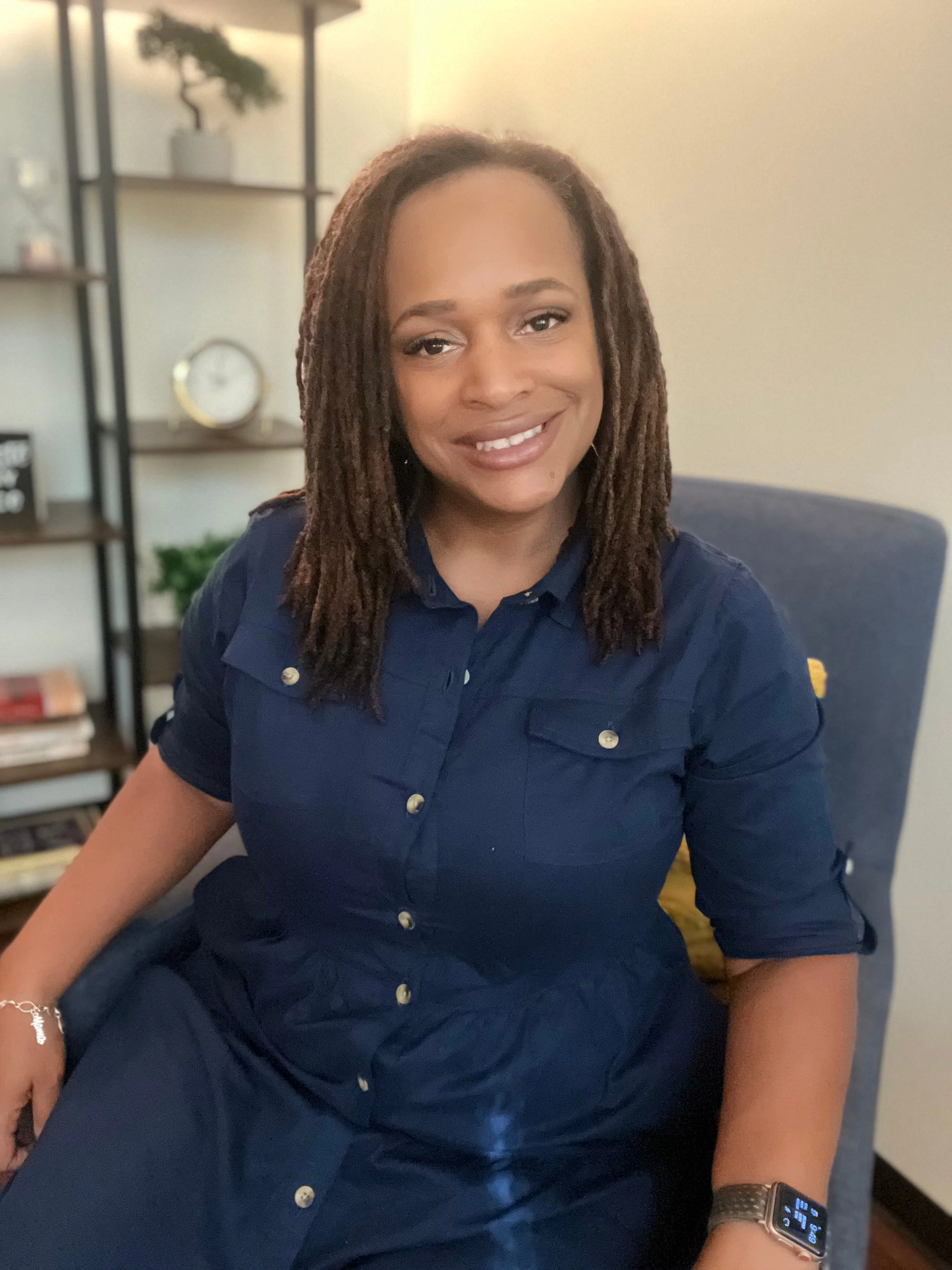 A woman with long, layered dreadlocks smiling while sitting indoors. She is wearing a dark blue button-up shirt, a silver bracelet, and a smart watch on her left wrist. In the background, there are shelves with decorative items, including potted plants, a clock, and books.
