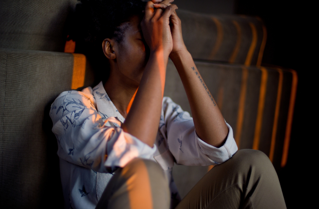 A woman sitting against a wall with her eyes closed, hands covering her face, appearing distressed or upset.