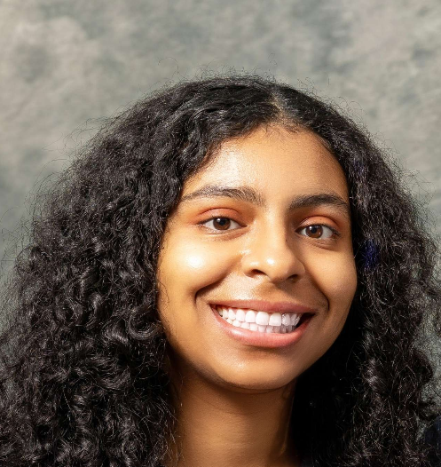 Portrait of a young woman with curly hair smiling against a gray background.