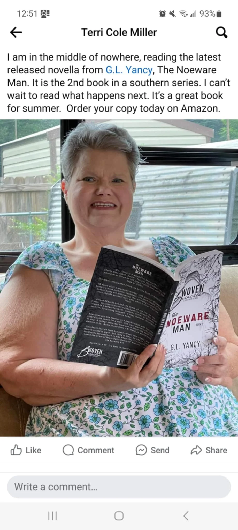 A woman with short gray hair, wearing a floral dress, sitting on a beige couch, holding a paperback book titled 'The Noeware Man' by G.L. Yancy, with a backyard visible through a window behind her.