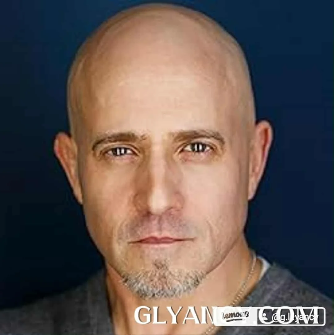 Close-up portrait of a bald man with light skin, blue eyes, and a goatee, wearing a gray shirt against a dark background.