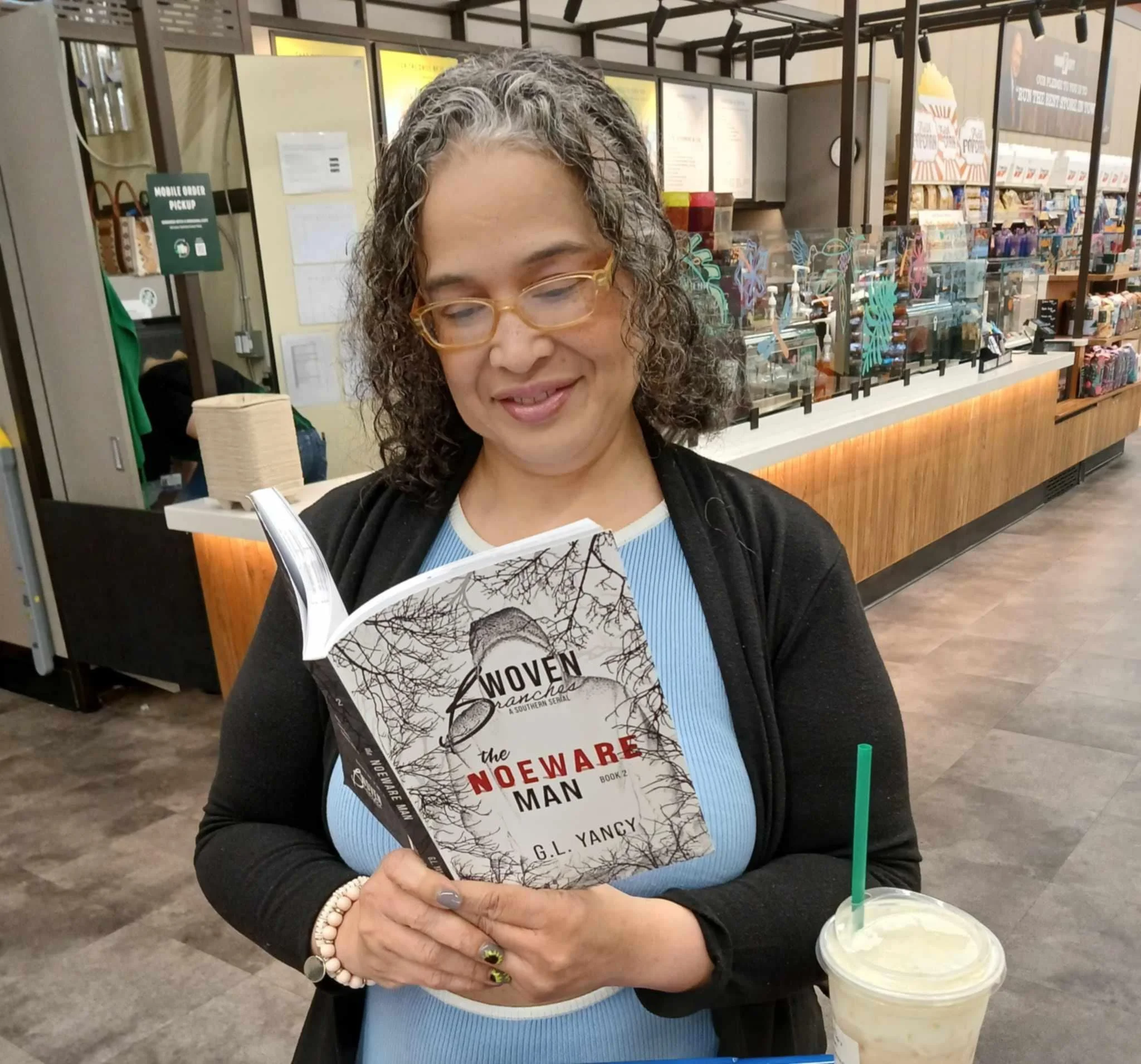A woman with glasses and curly hair smiling while holding a book titled 'The Noewearde Man' inside a bookstore or cafe, with a drink on the counter beside her.