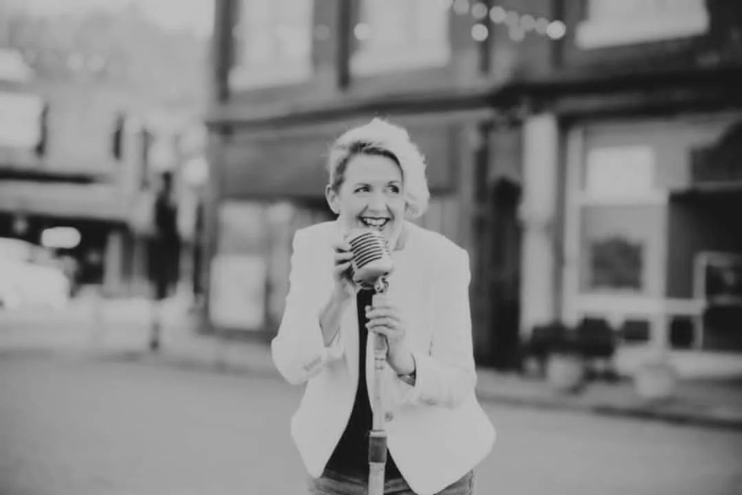 A woman with short, wavy hair smiling and holding a vintage microphone in a city street. She is dressed in a white blazer.