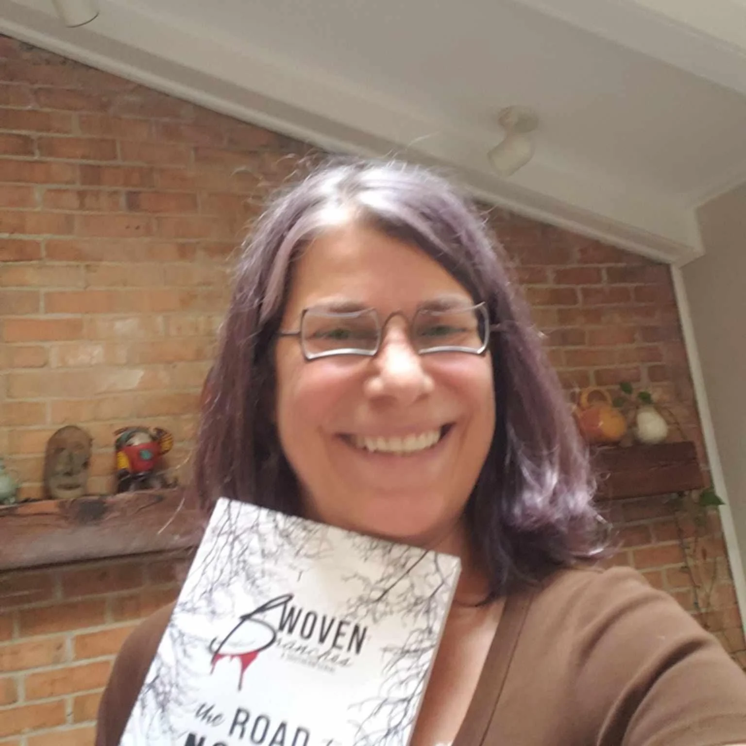 A woman with glasses and shoulder-length dark hair smiling and holding a brochure, standing inside a room with a brick wall and decorations on a shelf behind her.