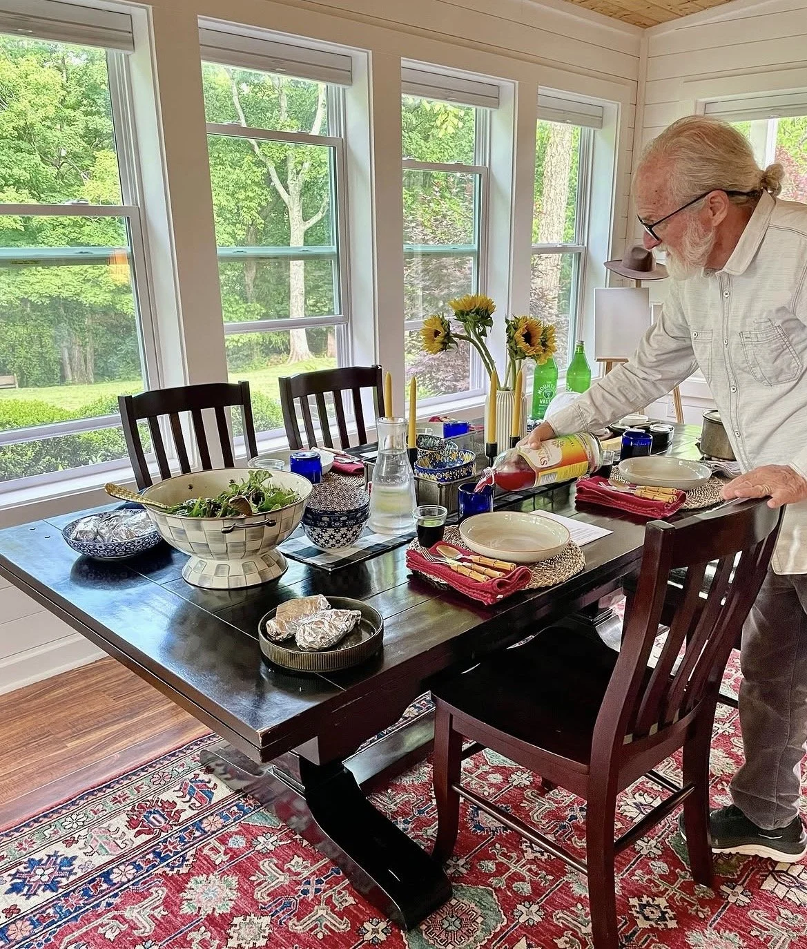 A man is preparing to serve wine at a dining table set for a meal in a sunlit room with large windows showing green trees outside. The table has a large salad bowl, wrapped baked potatoes, plates, cutlery, colorful glasses, and a vase with sunflowers.