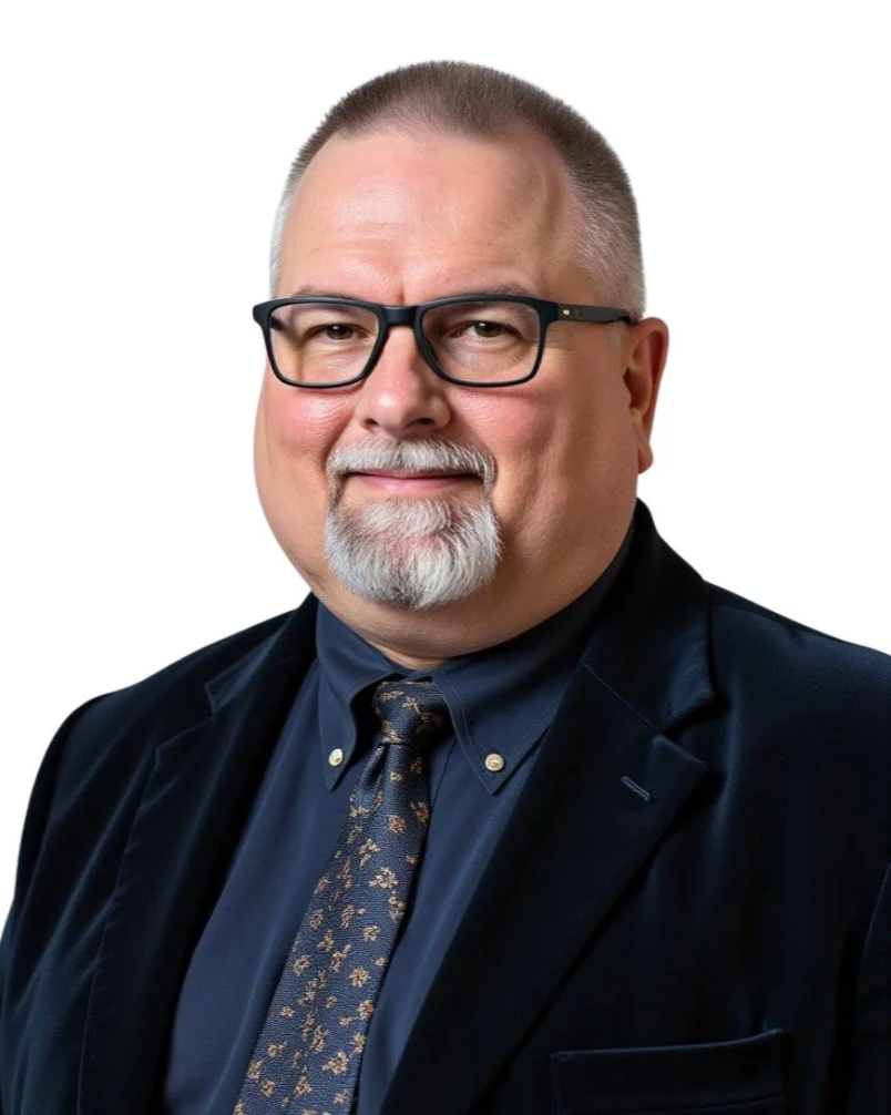 Portrait of a middle-aged man with glasses, a white beard, wearing a dark suit, dark blue shirt, and patterned tie, against a white background.