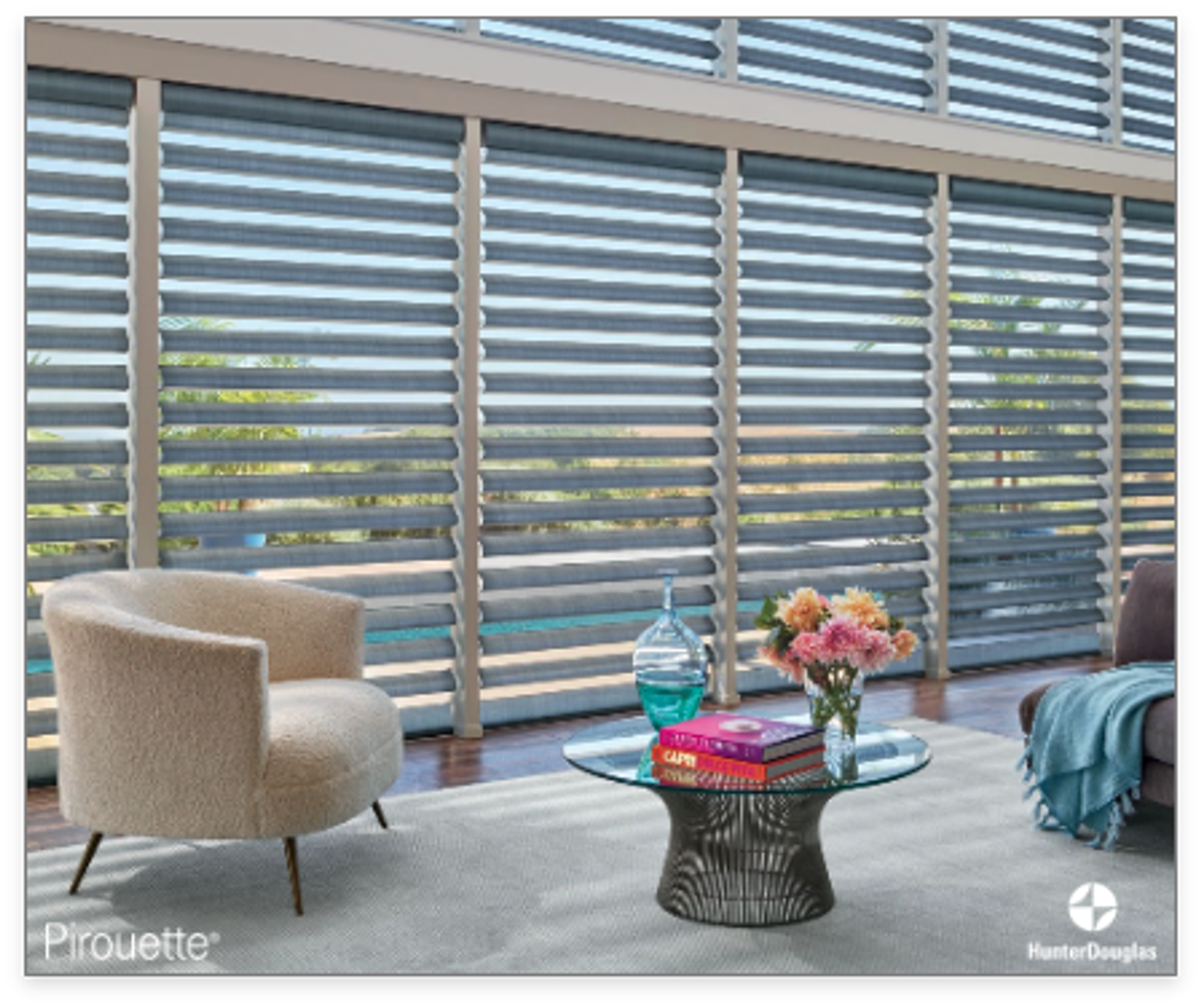 Contemporary living room with large horizontal slat window shades, a beige textured armchair, a round glass coffee table with a stack of books, a vase of colorful flowers, a blue blanket, and a rug on wooden flooring, overlooking an outdoor pool and greenery.