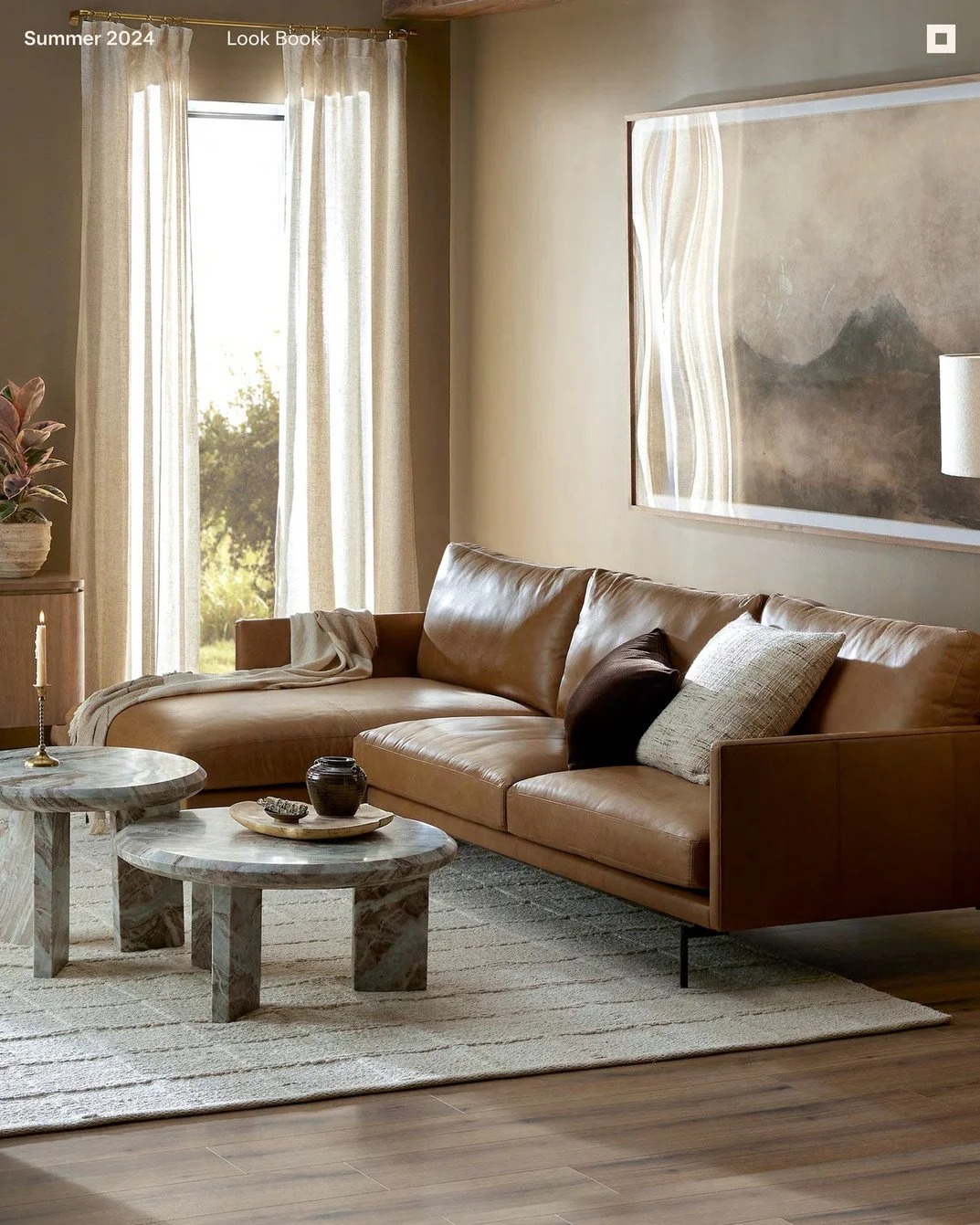 Living room with a tan leather sofa, two marble coffee tables, beige curtains, abstract wall art, decorative pillows, a candle, and a potted plant by the window