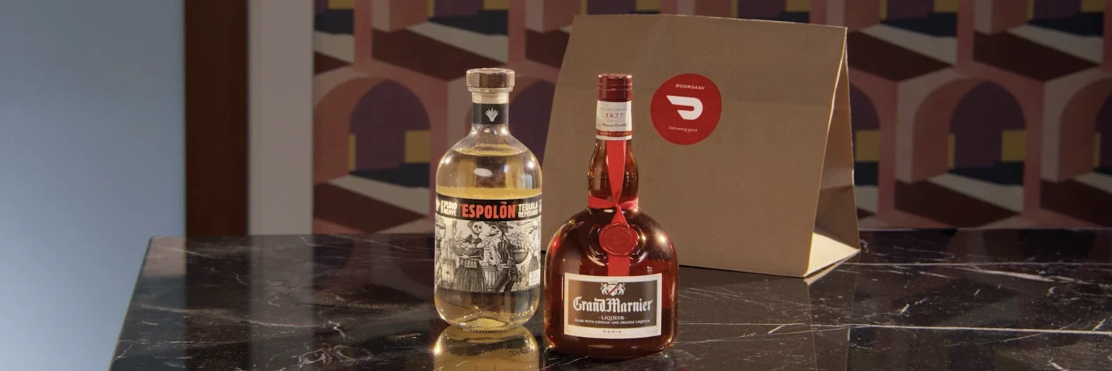 Two liquor bottles on a black marble table, with a cardboard box in the background. The left bottle is clear with a black, red, and white label, and the right bottle is red with a white label and a red seal. Production still from DoorDash commercial.