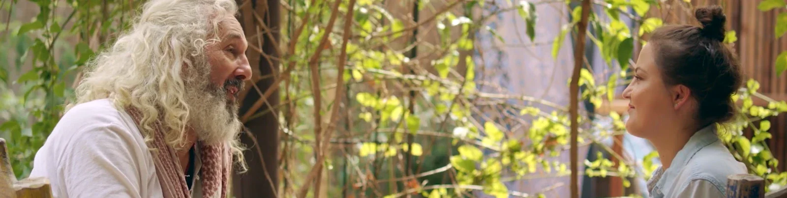 A man with long white hair and a beard smiling and talking to a woman with dark hair tied up, outside among green plants and trees. Production still from Lexus commercial.