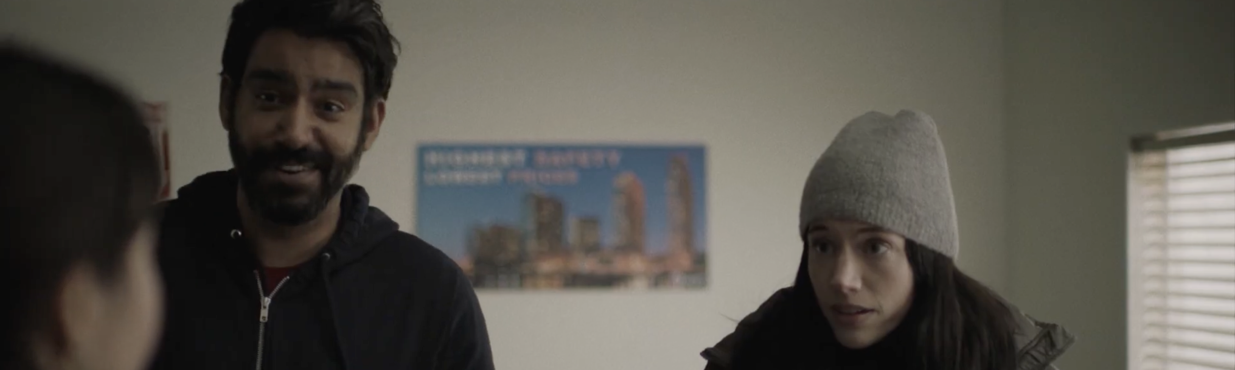 A man with a beard and dark hair, wearing a black hoodie, is talking to a woman with long dark hair, wearing a gray beanie and dark jacket, in a room with a window and a cityscape poster on the wall. A still from Next Exit
