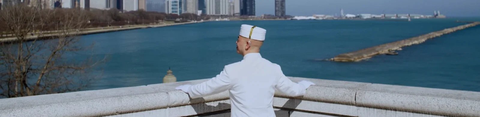 A man dressed in a white uniform and hat stands on a bridge, looking out over a body of water with a city skyline in the background. Production still from Peninsula Hotels commercial.