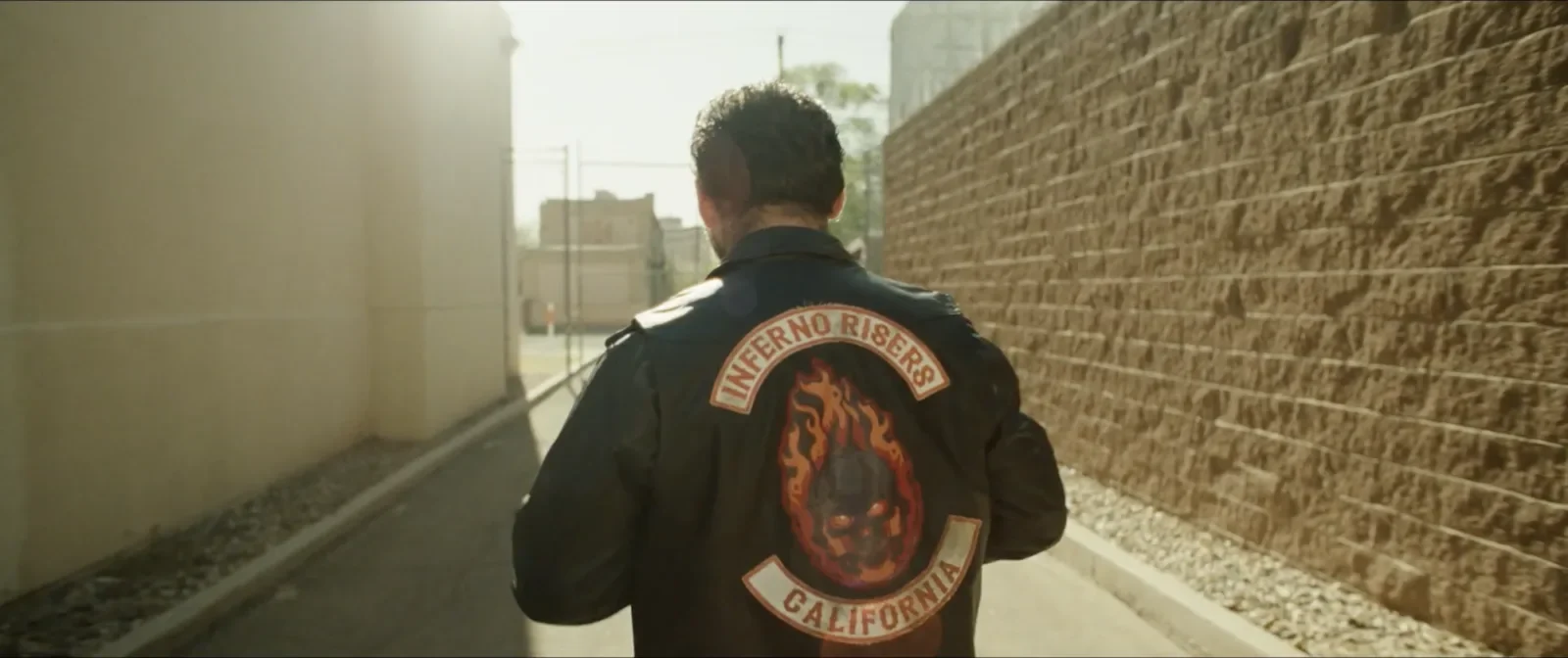 A man wearing a black jacket with a large patch on the back that says 'INFERNO RISERS CALIFORNIA' along with a fiery skull emblem. Production still from On The Run.