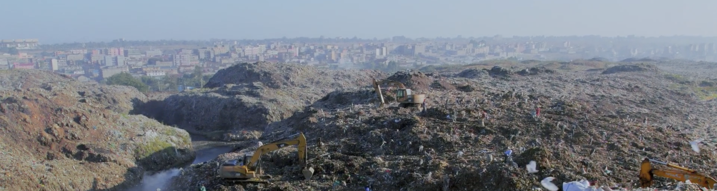 A landfill with multiple excavators working on piles of trash. In the background, a city skyline with numerous buildings can be seen. A still from Earthbound.