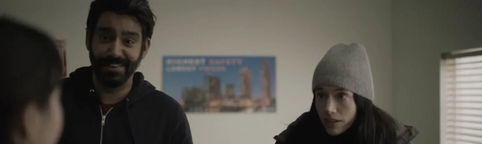 Three people talking in an indoors setting with a cityscape poster on the wall and a partially visible window with blinds. Production still from Next Exit.