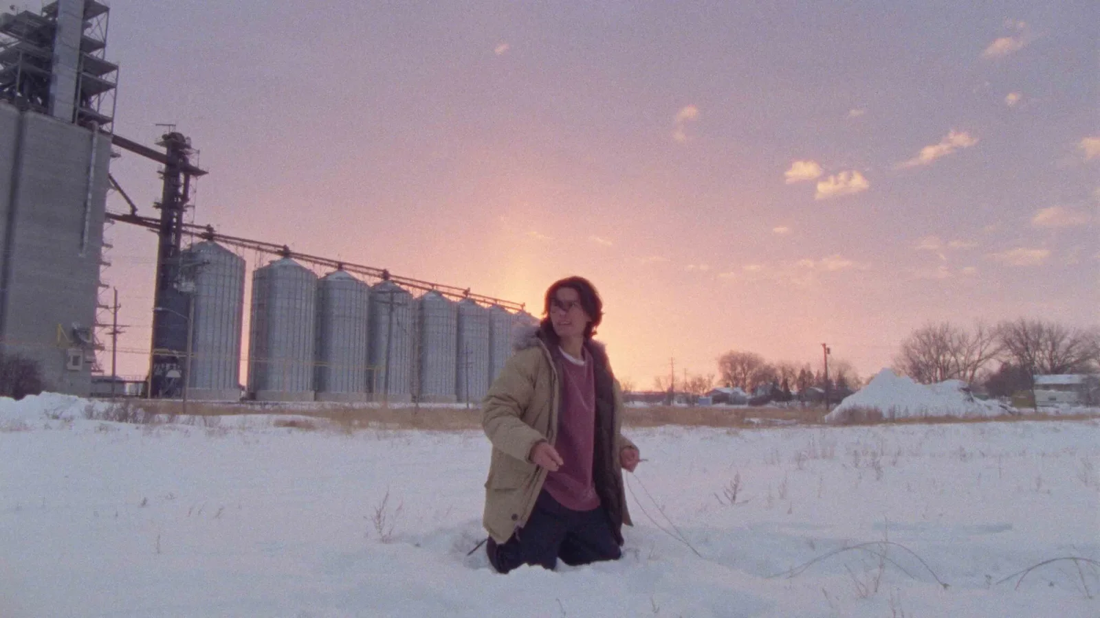 Person kneeling in snow during sunset with grain silos and trees in the background. Production still from How To Blow Up A Pipeline