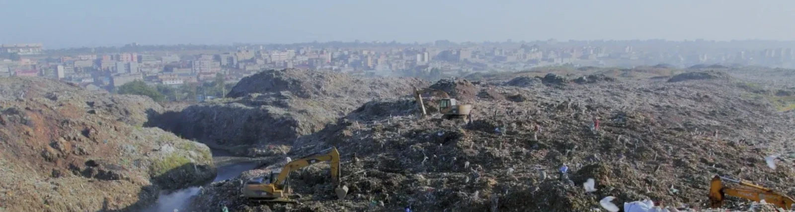 A large landfill with multiple bulldozers and excavators working on piles of trash, overlooking a city in the background. Production still from Earthbound.