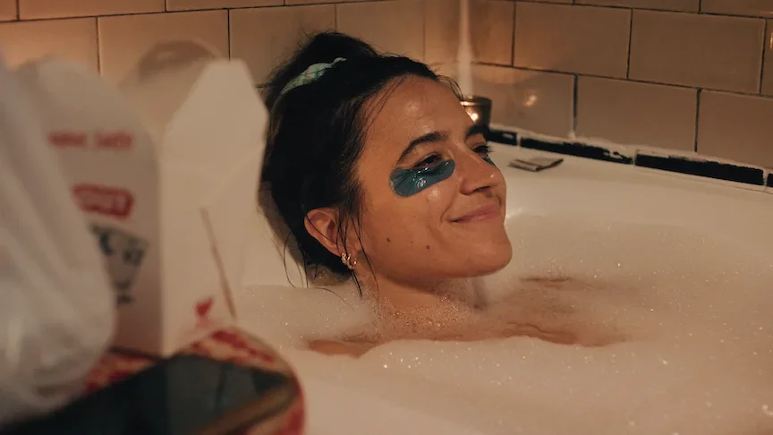 Woman relaxing in a bubbling bathtub with face mask on, enjoying a moment of self-care. Production still from Homebodies.