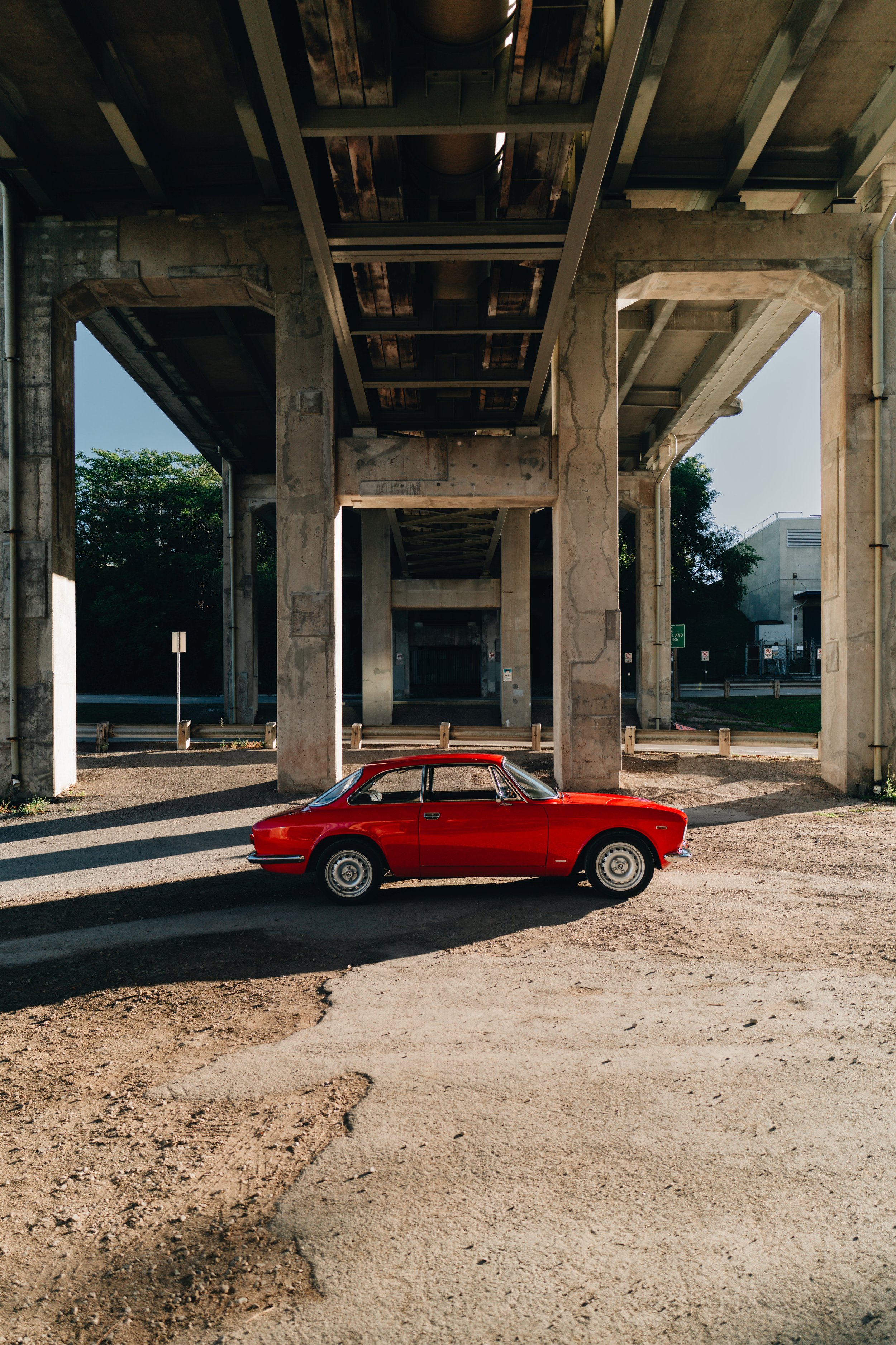 A red classic car parked under an overpass with concrete pillars and a highway above, trees and a building visible in the background.