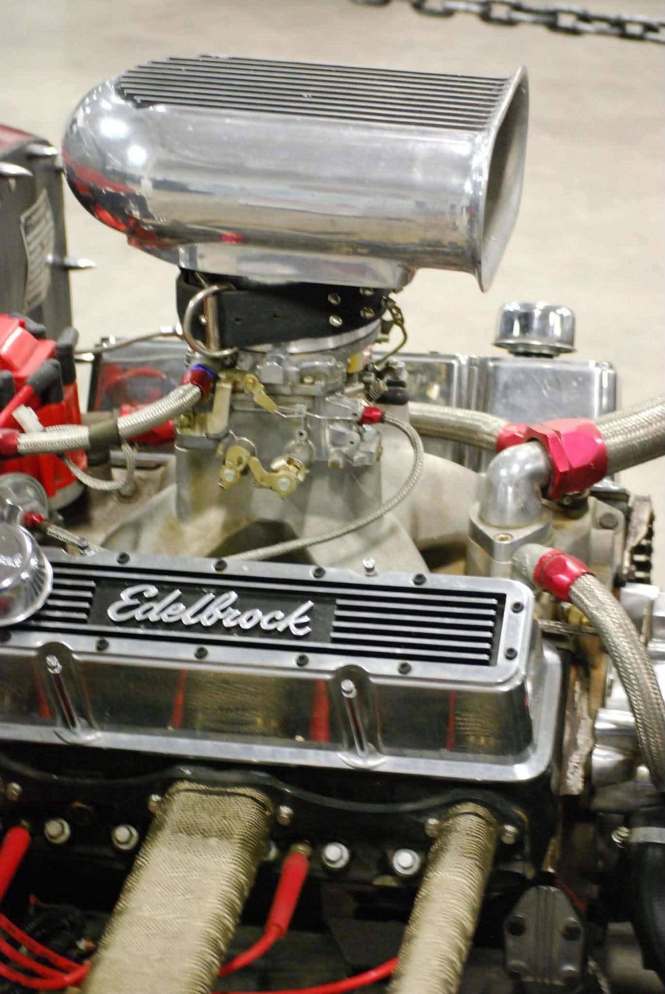 Close-up of a vintage Edelbrock carburetor with a chrome air scoop mounted on a racing engine, with various hoses and metallic components visible.