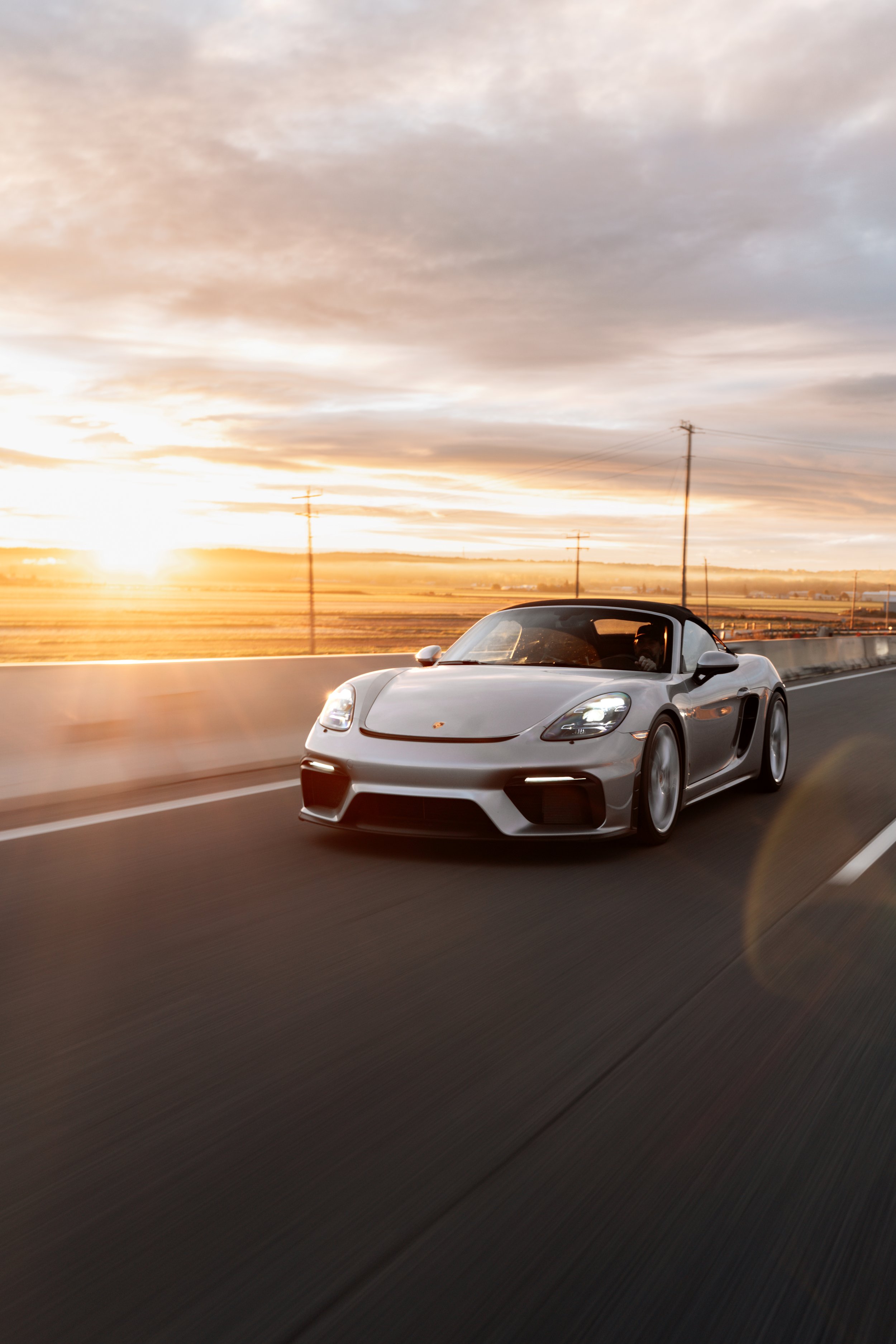 A silver sports car driving on a highway during sunset with a cloudy sky and power lines in the background.
