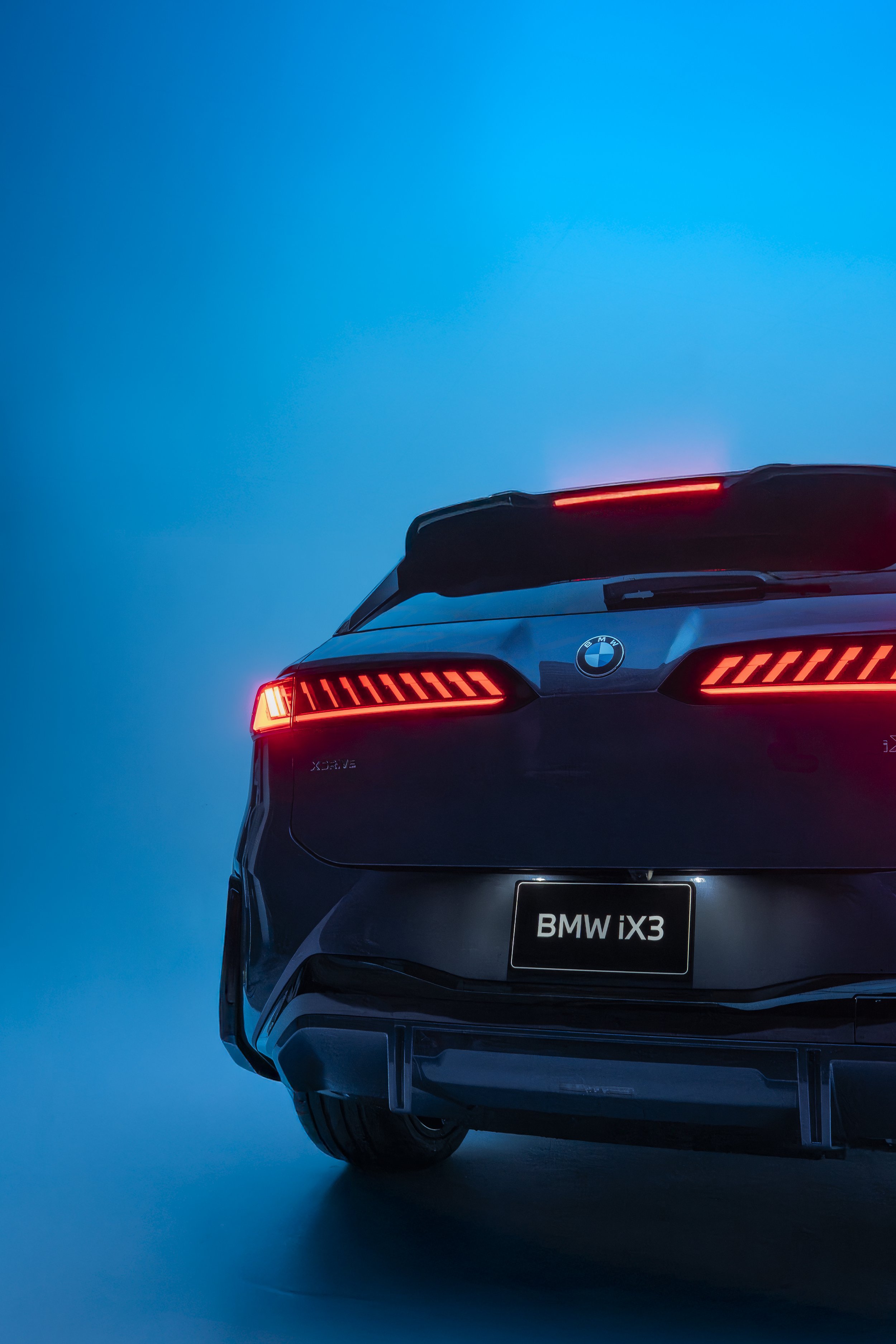 The rear view of a black BMW iX3 electric SUV with red tail lights illuminated and a blue gradient background.