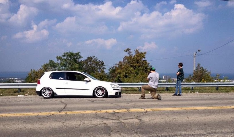 Two people pull over on the side of a road with a lowered white car parked nearby. One person is kneeling and taking a photo, while the other stands next to the guardrail. Trees and a city skyline are in the background under a partly cloudy sky.