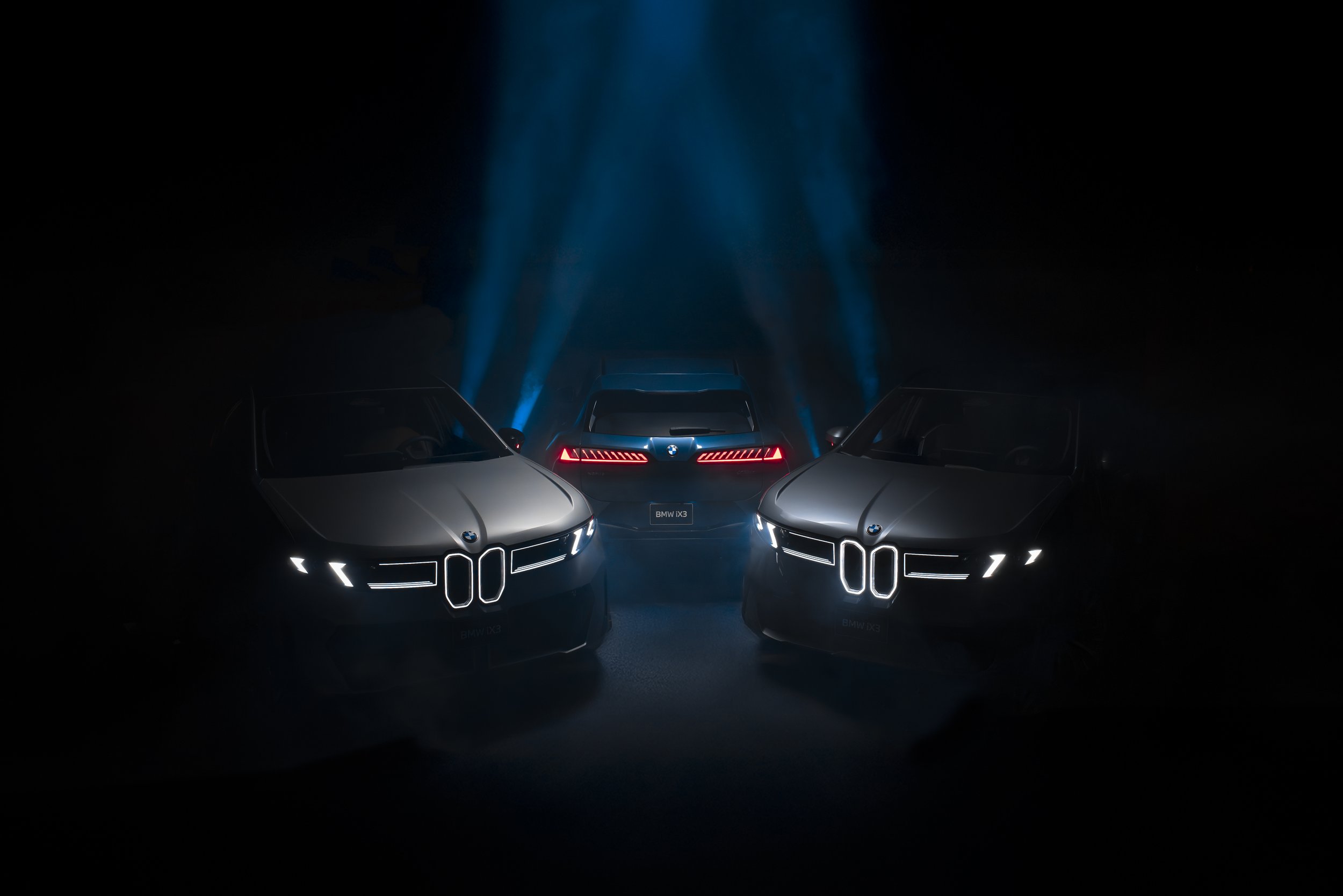 Three futuristic cars with illuminated front grilles and taillights are in a dark setting, with the central car displaying a BMW logo and model badge.