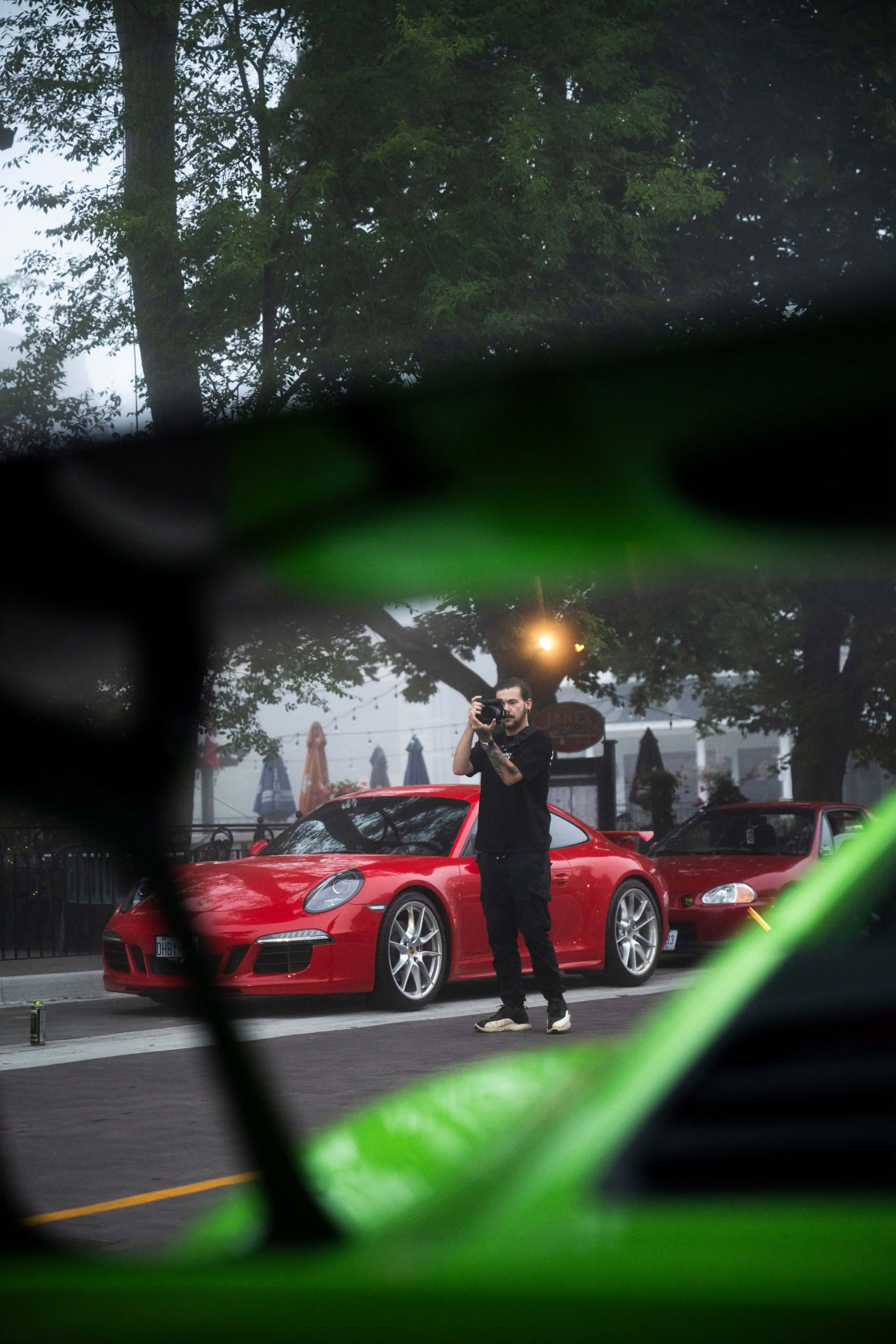 A man taking a photo with a camera in front of a red sports car parked on the street, with trees and umbrellas visible in the background, seen through a green metallic structure.