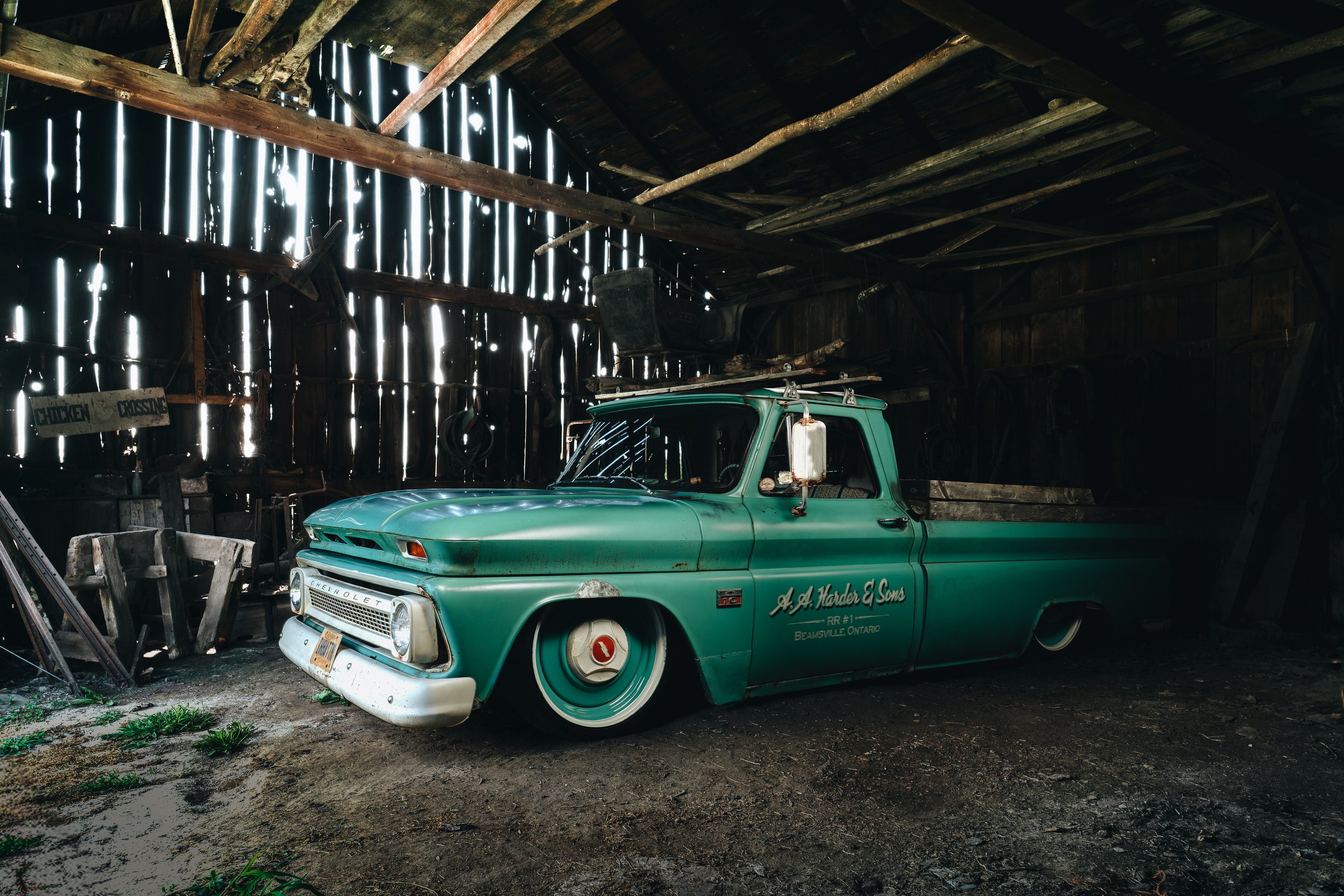 An old green Chevrolet pickup truck with a white front bumper and stylized text on the side, parked inside a rustic wooden barn with scattered tools and signs, including a chicken crossing sign.