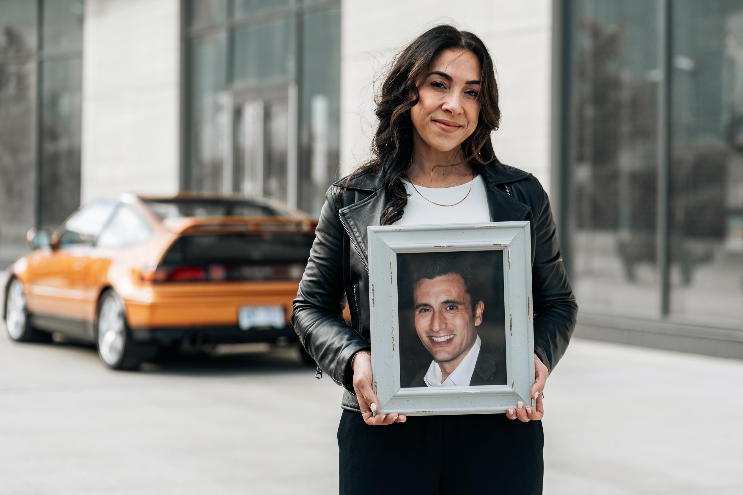 A woman with dark wavy hair, wearing a leather jacket and white shirt, holding a framed photograph of a smiling man in front of a city street with a yellow taxi in the background.