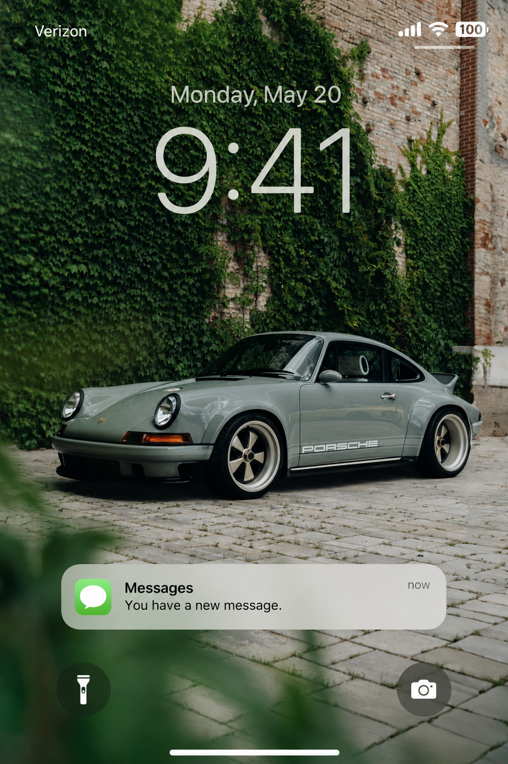 iPhone lock screen with the time 9:41, date Monday, May 20, over a background of a vintage gray Porsche 911 parked on a cobblestone street against a brick wall covered with green ivy.