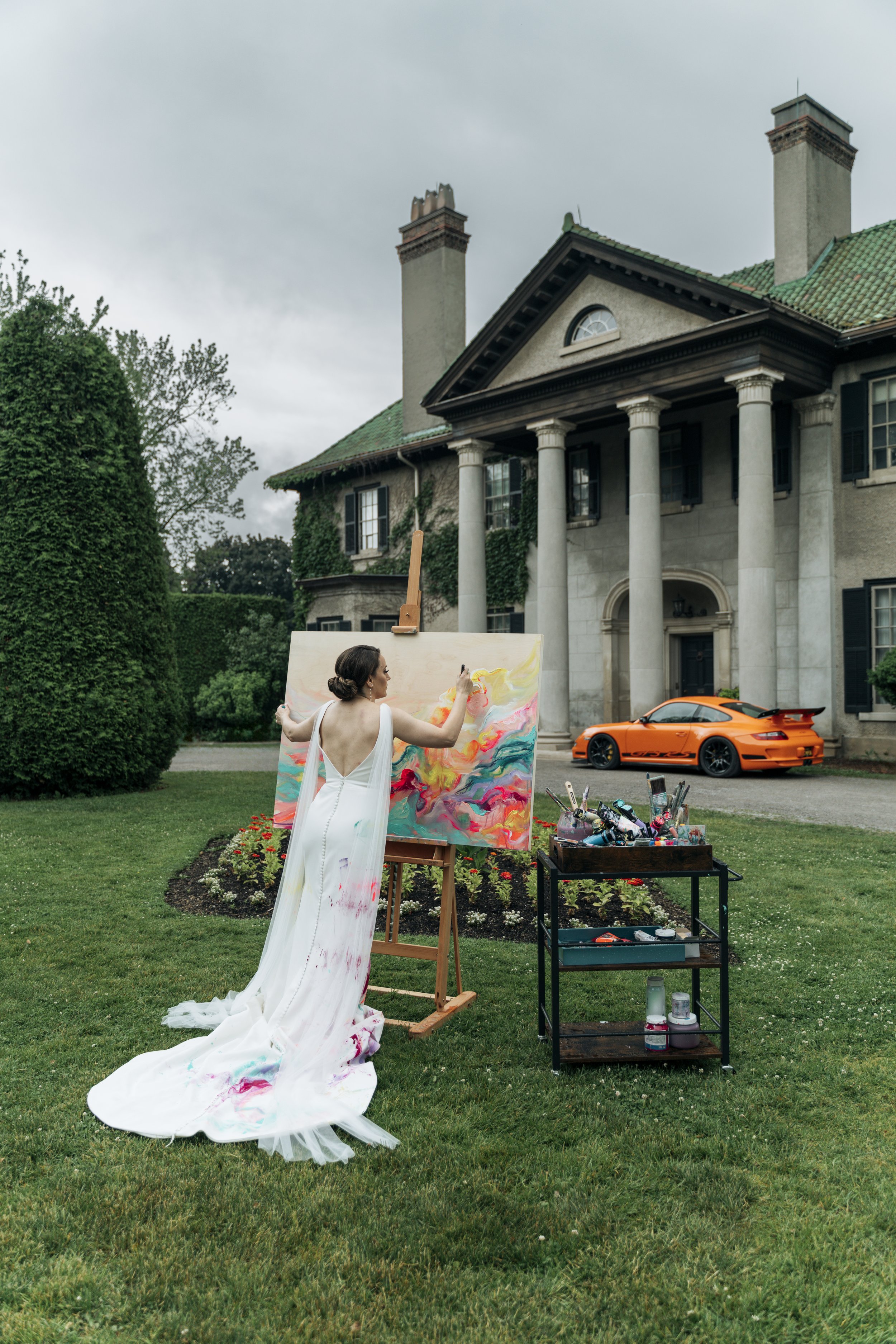 A woman in a white dress with colorful paint marks on the hem creating art on a large canvas outdoors in front of a mansion with columns, chimneys, and ivy-covered walls. There is an orange sports car parked on the driveway and art supplies on a nearby cart.