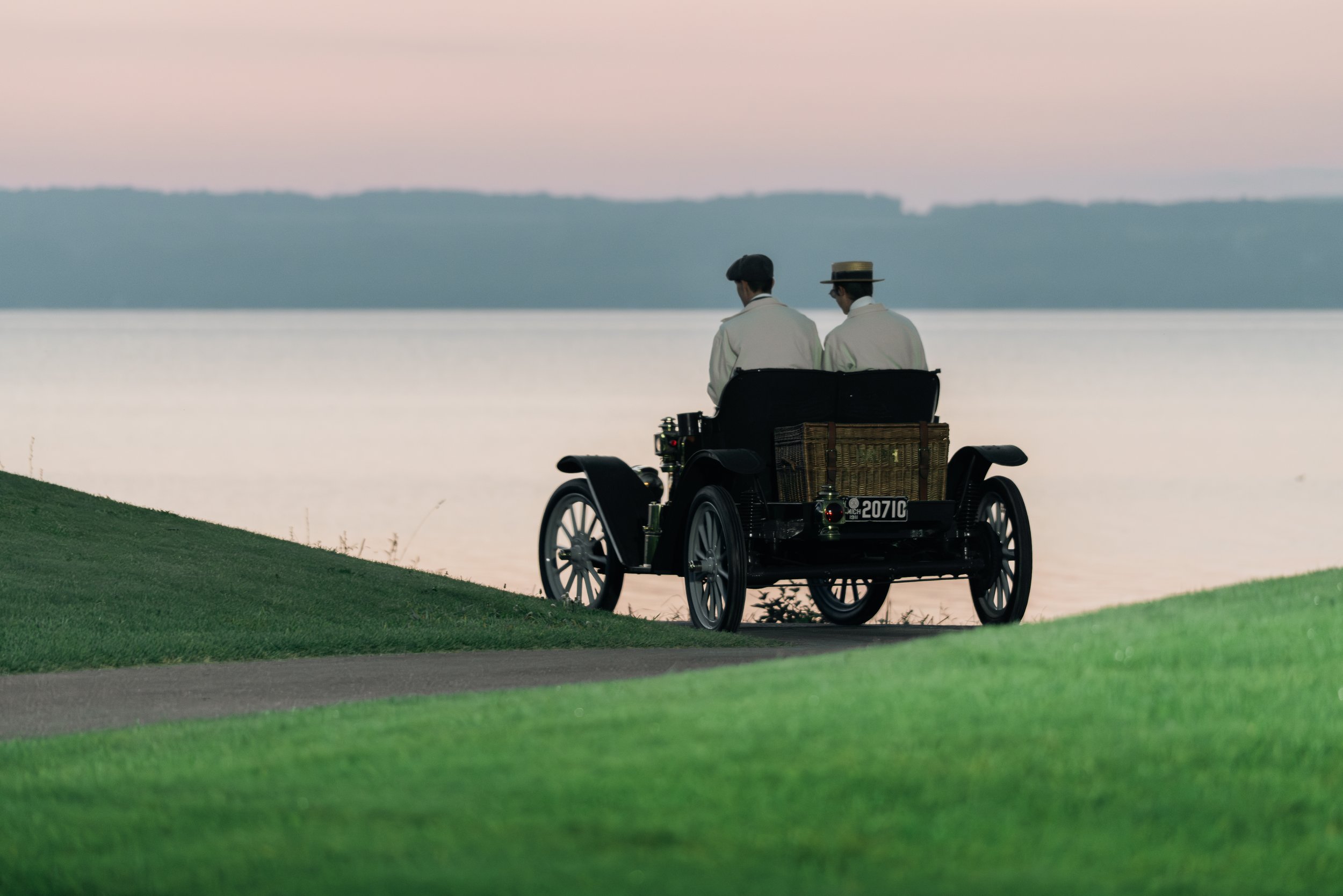 Two people in vintage attire sitting in a picnic carriage near a lake during dusk, with a calm body of water and distant hills in the background.