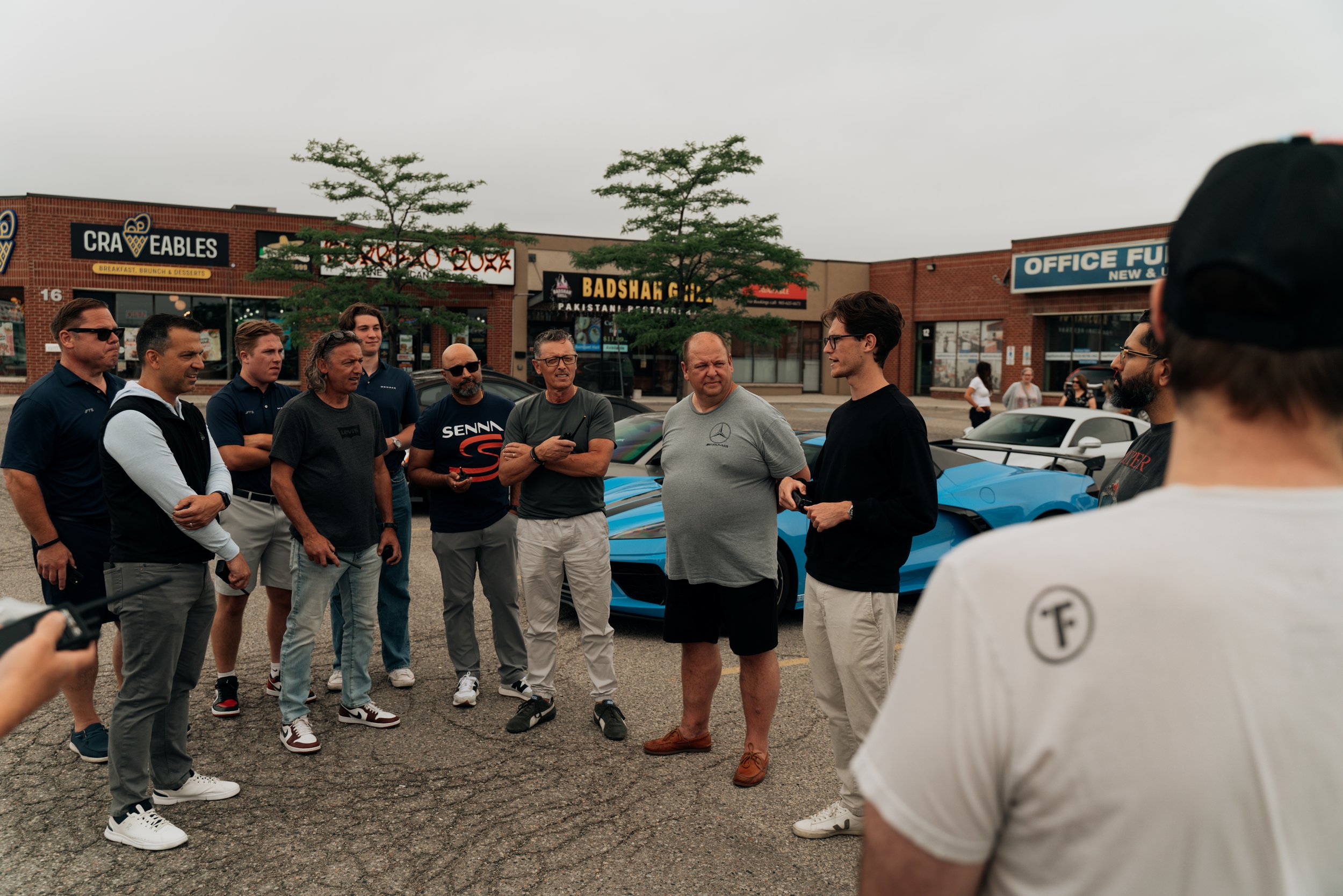 A group of men standing outside in a parking lot, engaged in conversation, with storefronts behind them including a restaurant and an office furniture store, under an overcast sky.