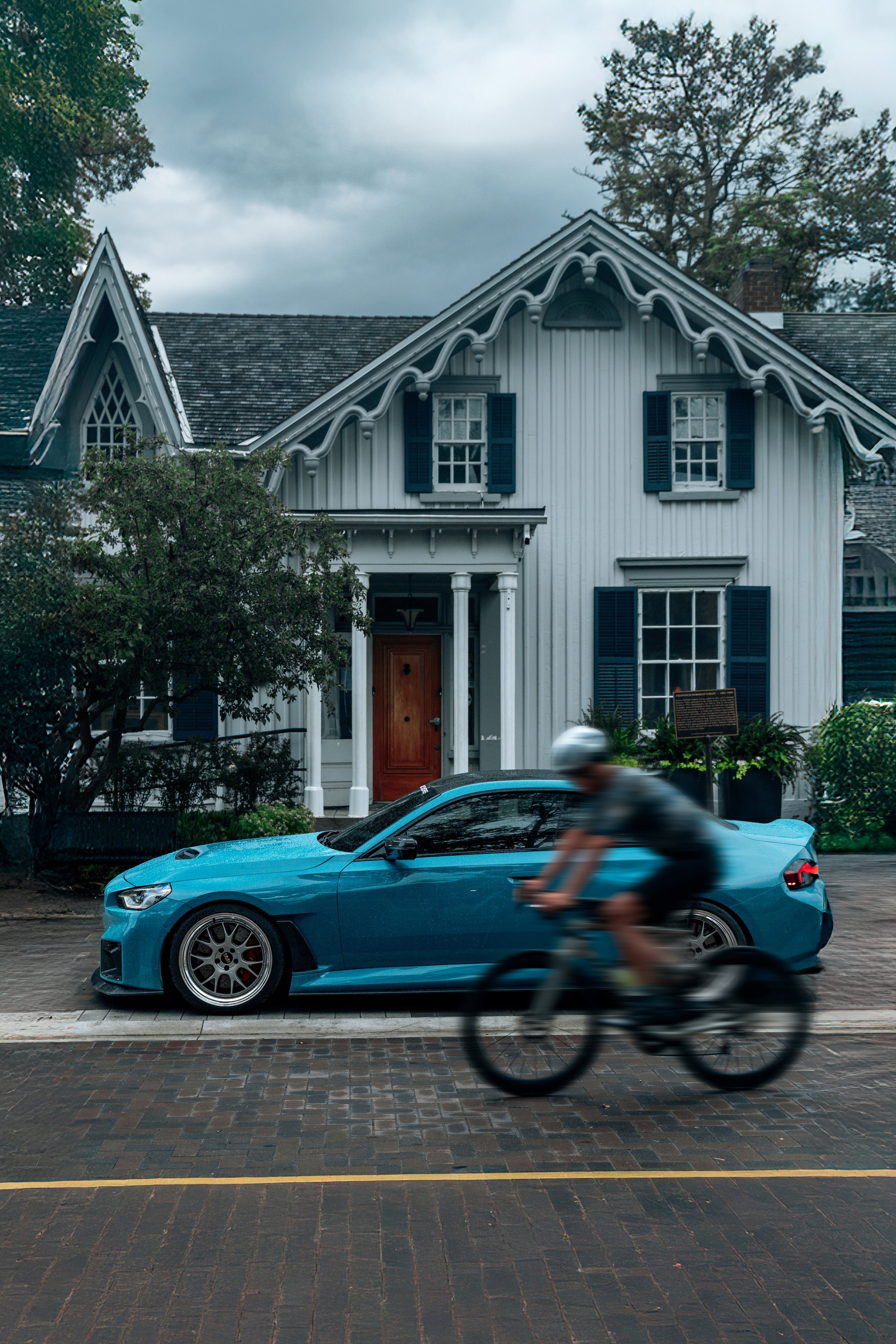 A blue sports car parked on a brick street in front of a white house with dark shutters and a porch. A person in a gray shirt and helmet is cycling past the car, appearing blurred due to motion.